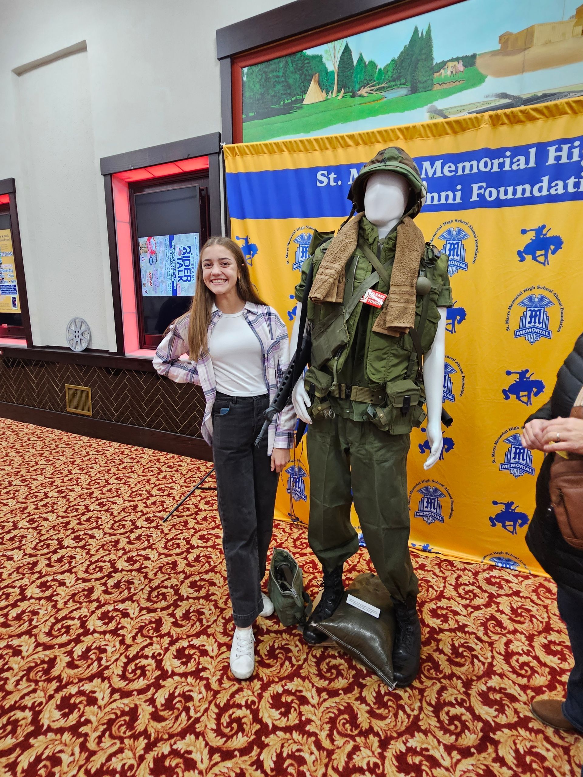 Young person poses with a military uniform display; indoor setting with banner and patterned carpet.