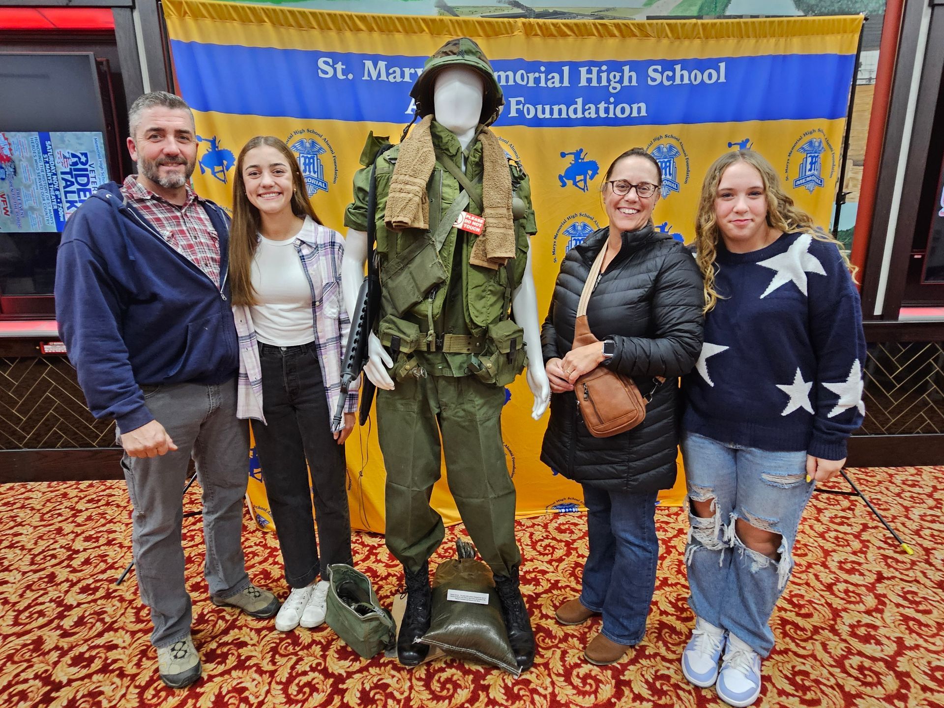 Three men stand beside a military uniform display. Background features a banner for St. Marys Memorial High School Foundation.