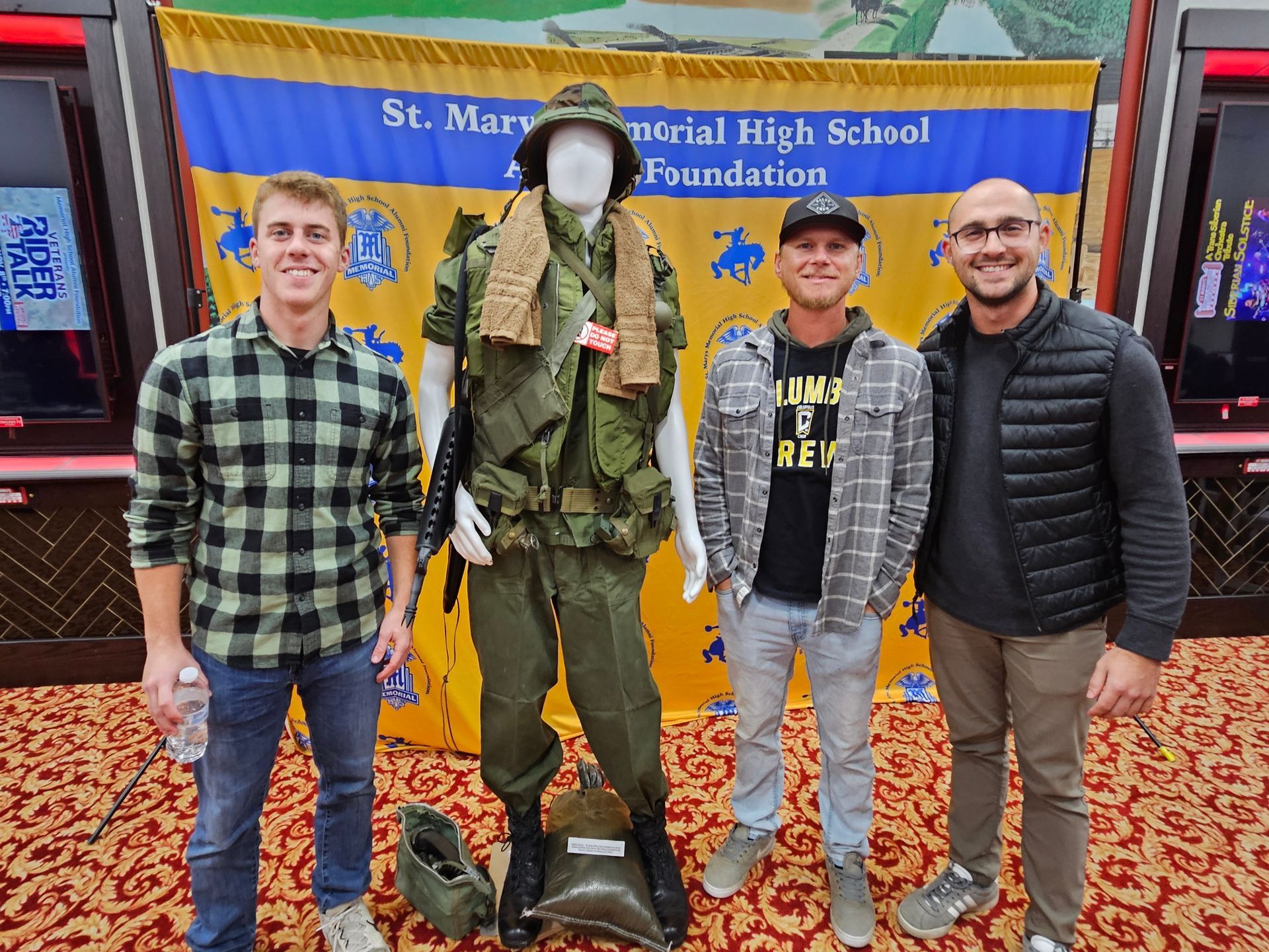 Two men stand by a mannequin in a military uniform. Background: St. Mary's school banner.
