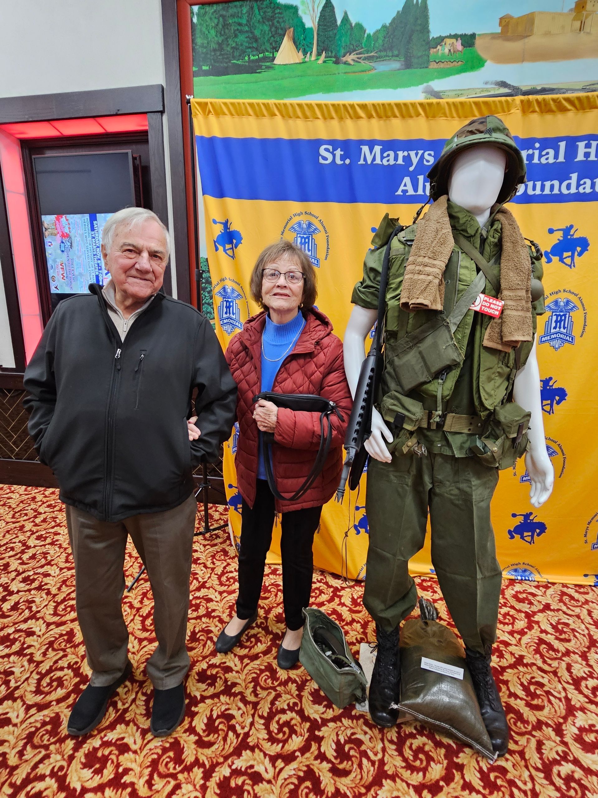 Two people stand near a mannequin wearing a military uniform. St. Mary's Memorial Hall banner in background.
