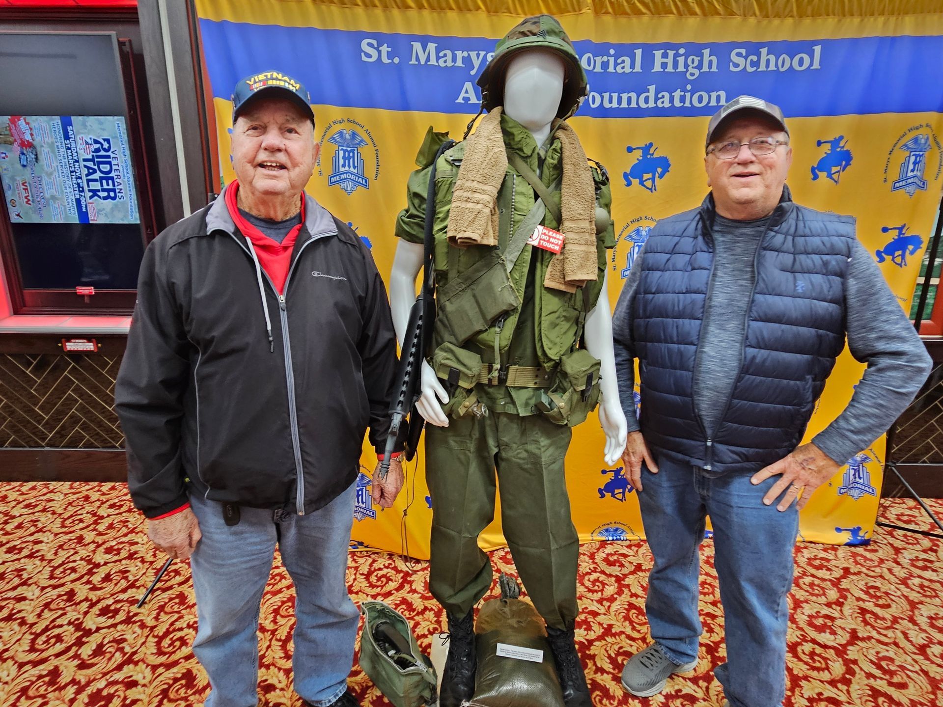 Two men stand by a mannequin in a military uniform. Background is a school banner.