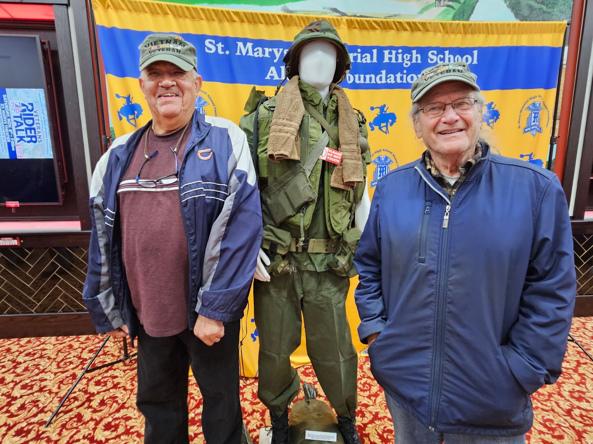 Two men stand next to a mannequin wearing a military uniform in front of a school banner.