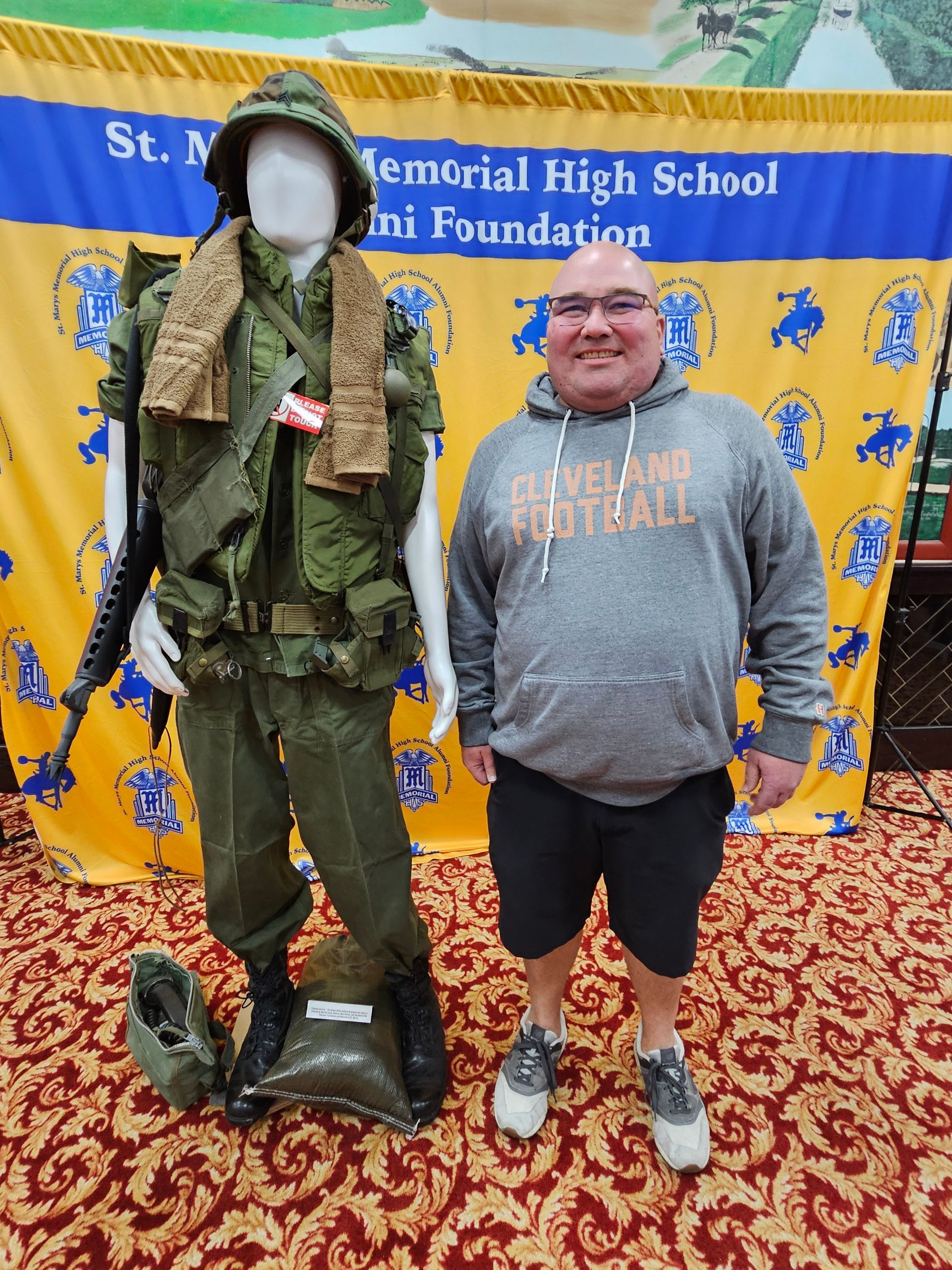 Man poses next to a mannequin dressed in a military uniform. Background: banner, rug.