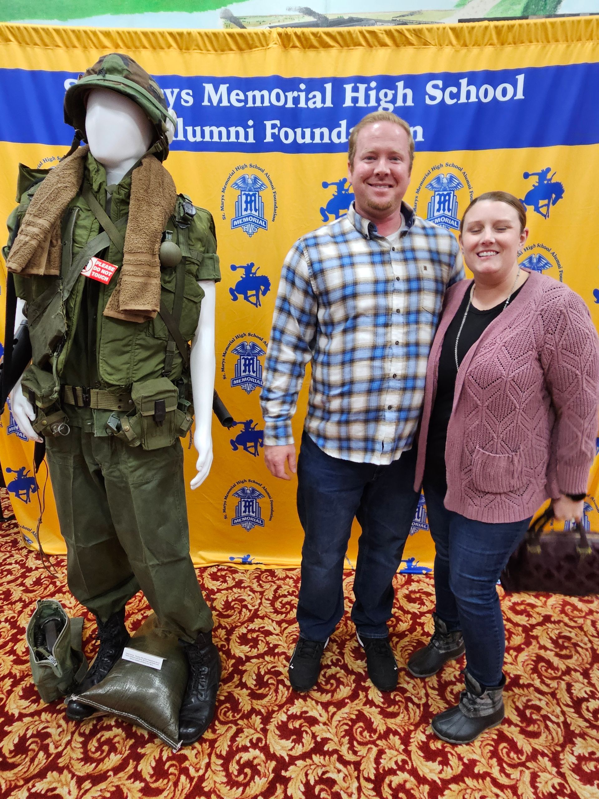 Man and woman standing beside a mannequin in a military uniform in front of a banner.