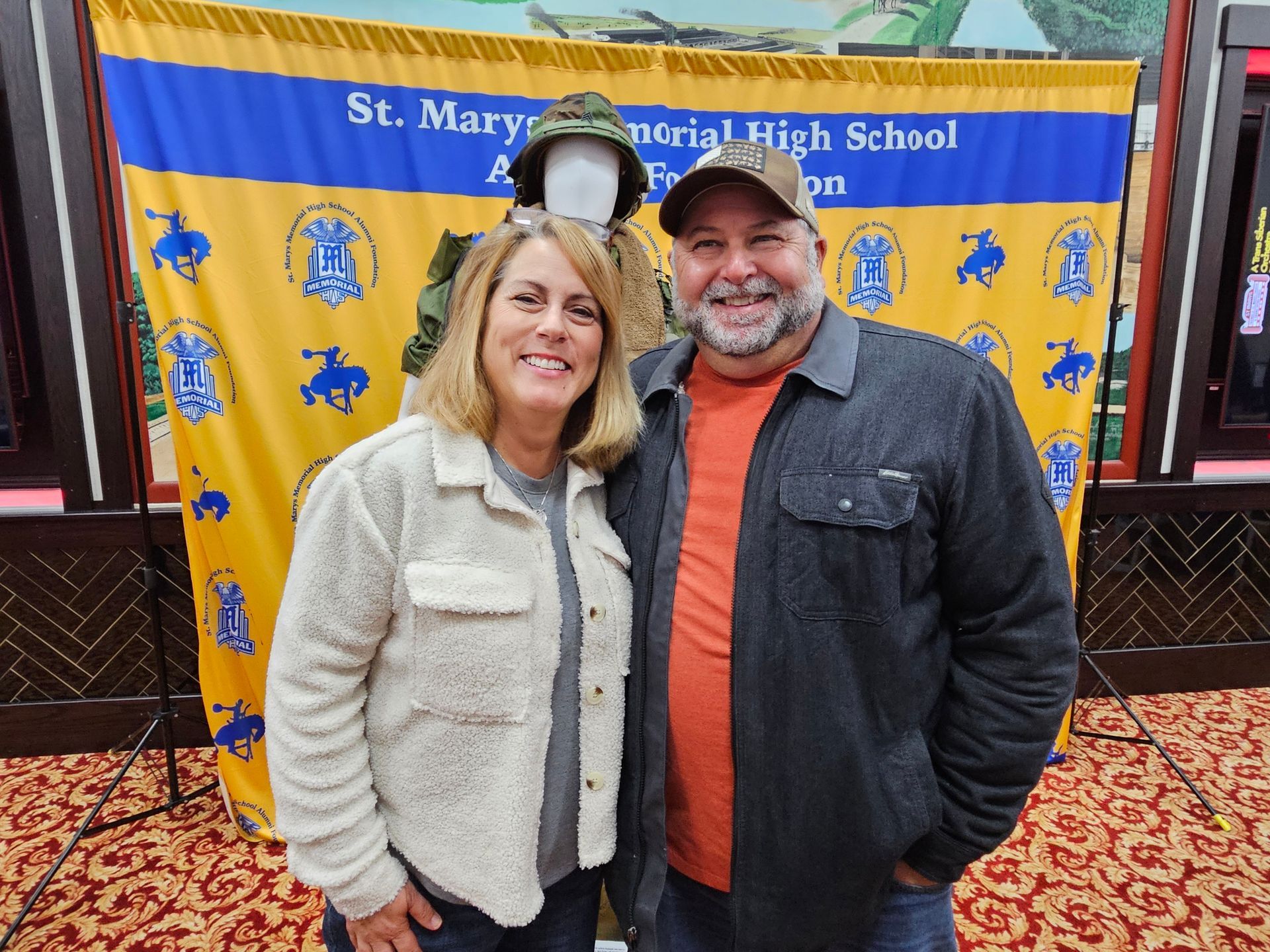 Couple poses for a photo in front of a banner for St. Marys Memorial High School, a mannequin in military uniform.
