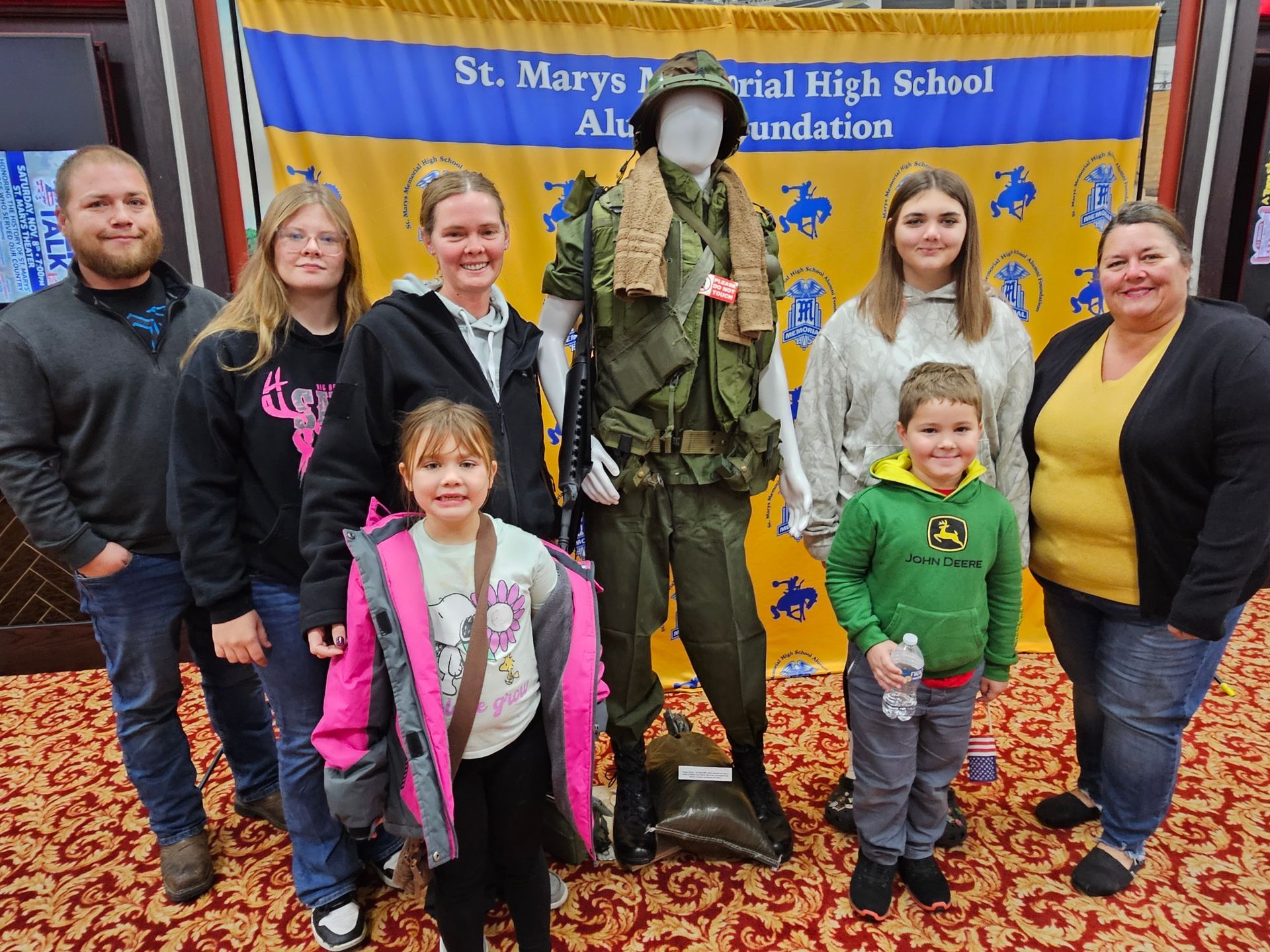 A group poses with a mannequin in military uniform, in front of a banner. Carpeted setting.