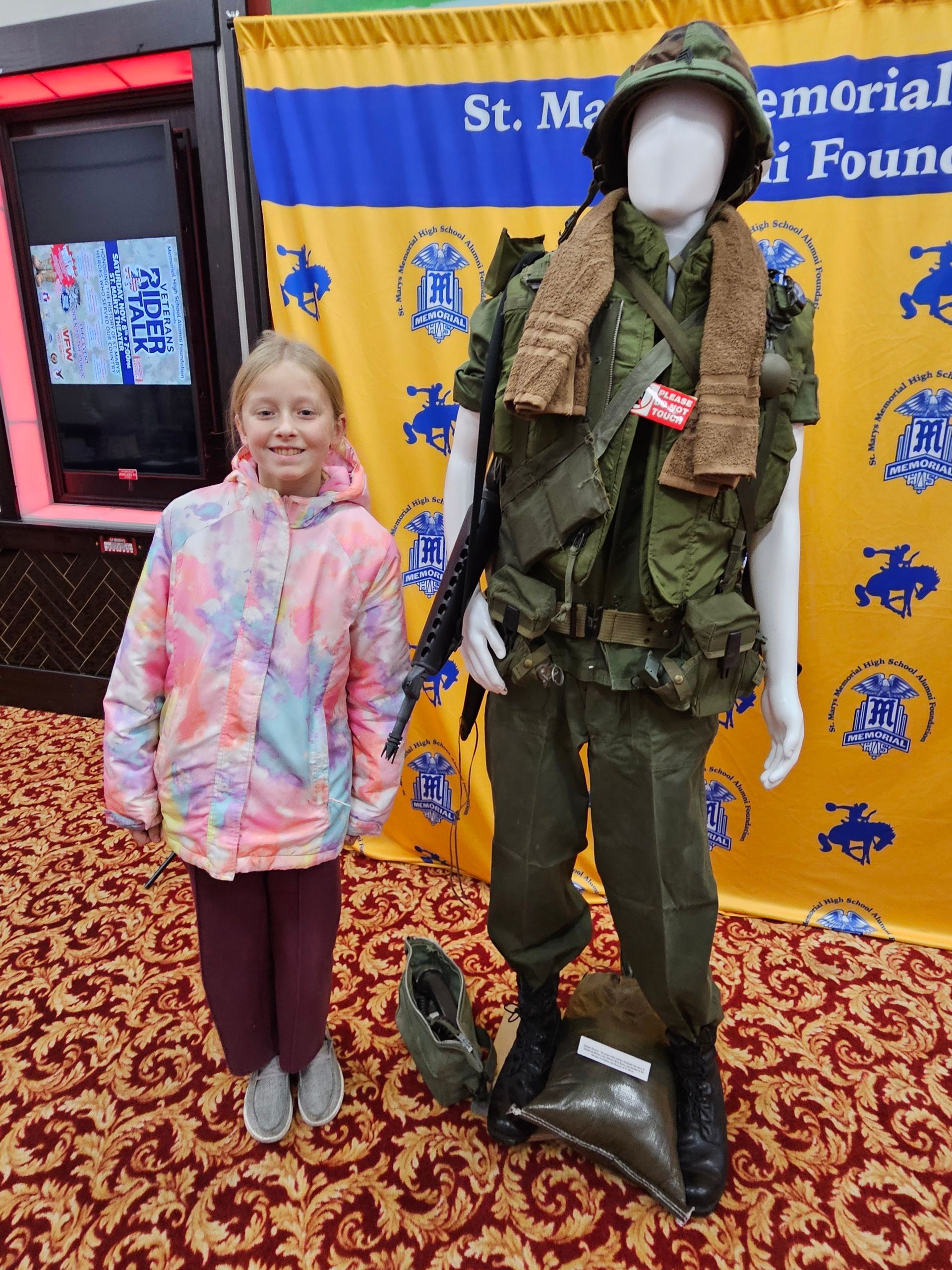 Girl stands next to a mannequin dressed in military fatigues, in front of a banner.