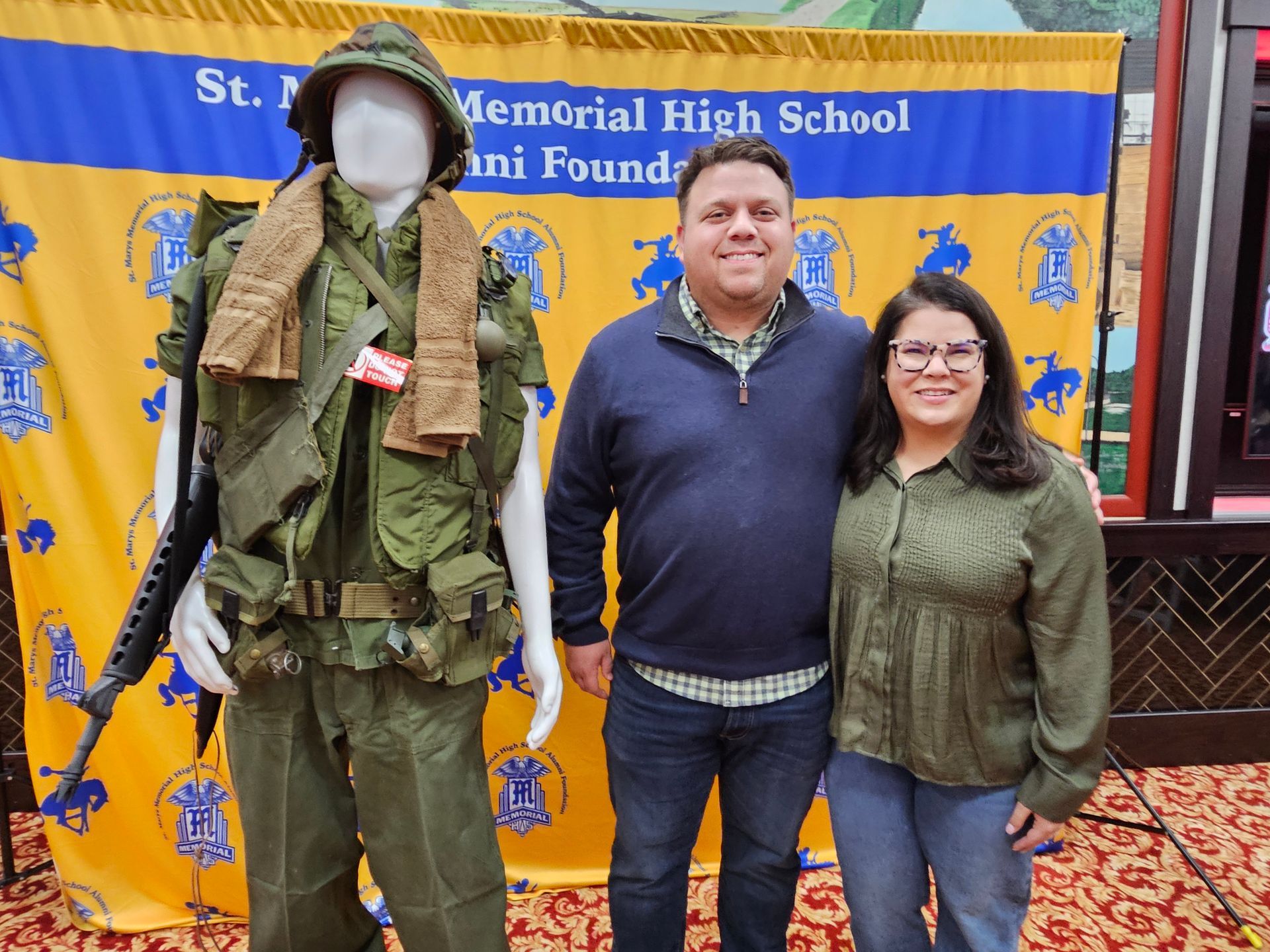 Man and woman smiling by a mannequin in military gear. A banner for St. Mary's High School is behind them.