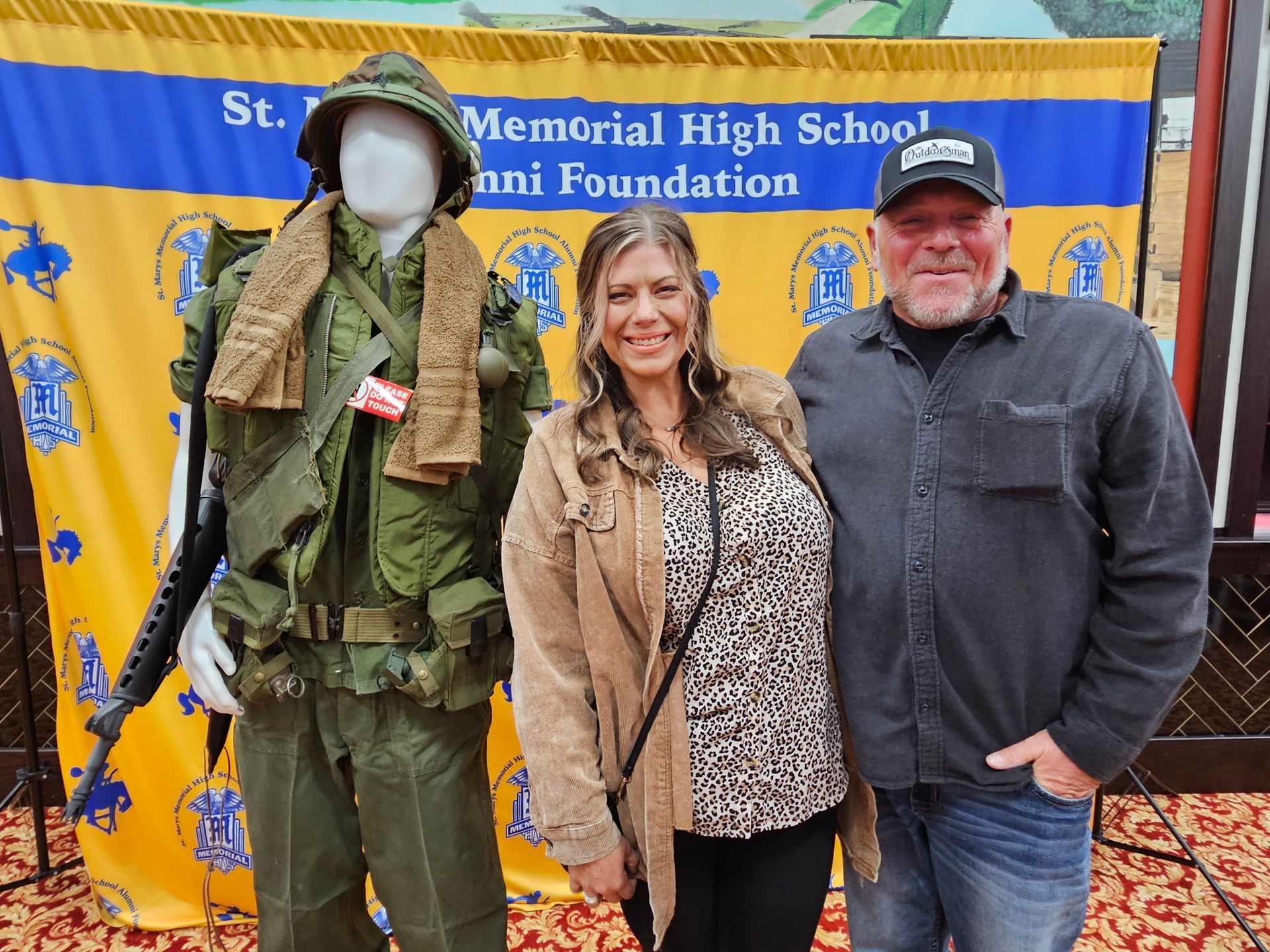 Man and woman stand next to a mannequin in a military uniform. Behind them is a school banner.