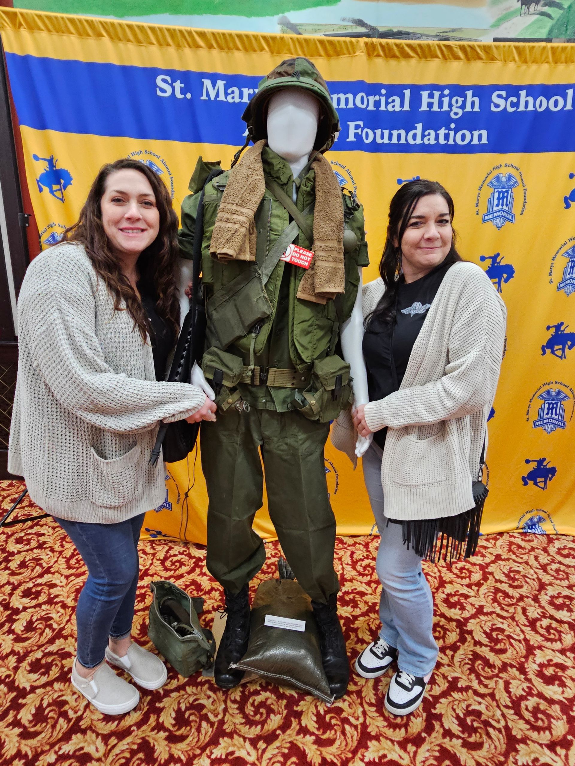 Two women beside military uniform display. Gold and blue backdrop, patterned floor.