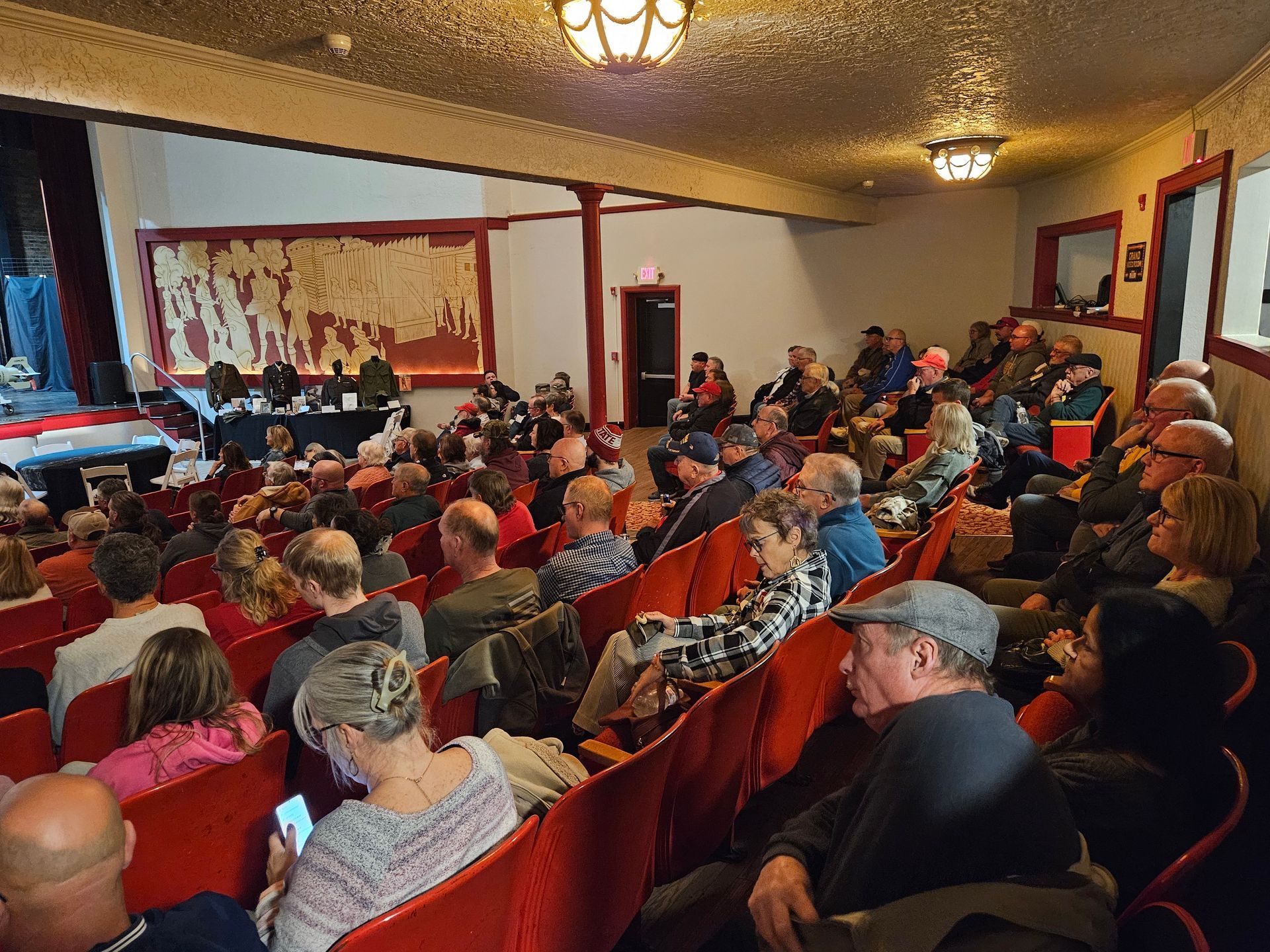 Audience seated in a red-seated theater, watching a presentation on stage.