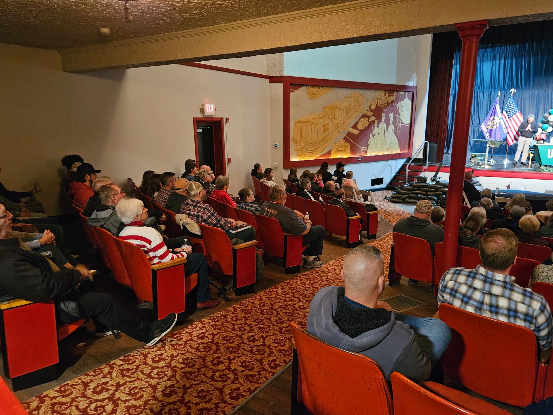 Audience watching a performance on stage in a theater with red seating and gold trim.