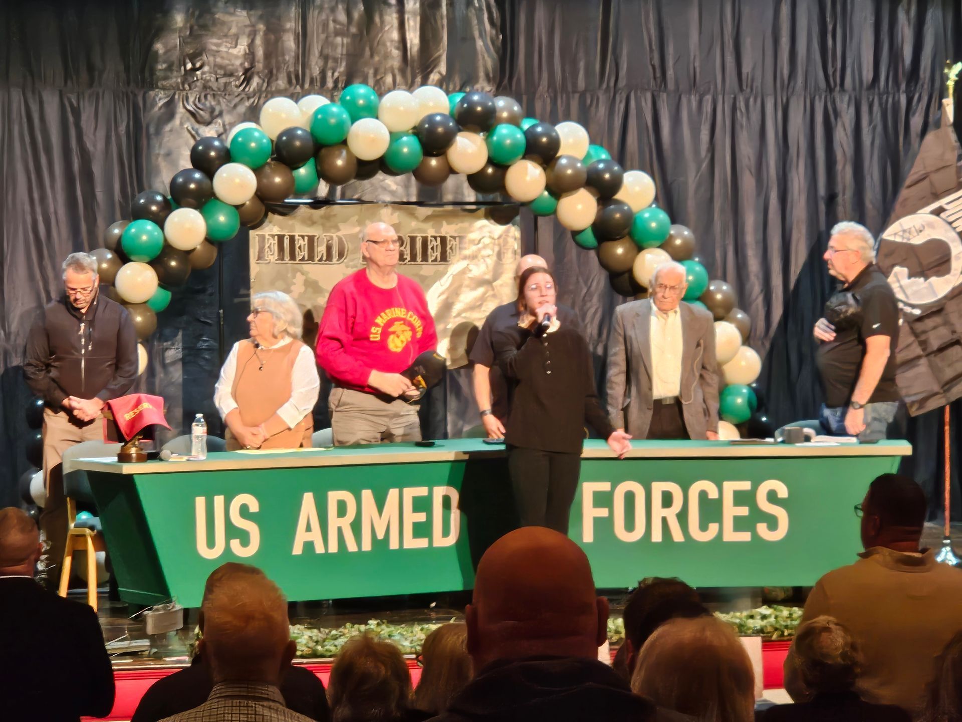 People on a stage with a US Armed Forces banner and a balloon arch, speaking at an event.
