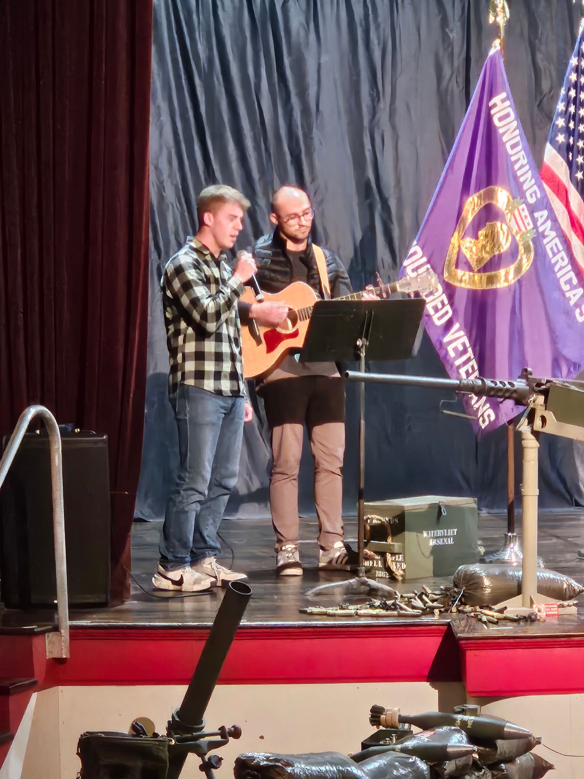 Two men on stage singing, one with a microphone, guitar. Flags and military items in the background.