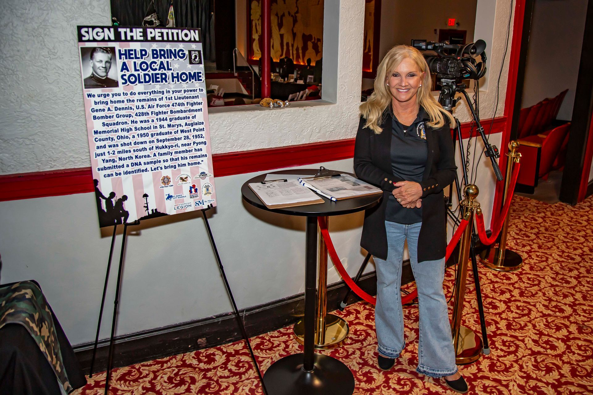 Woman standing by a petition sign, red rope and stanchion, camera on tripod.