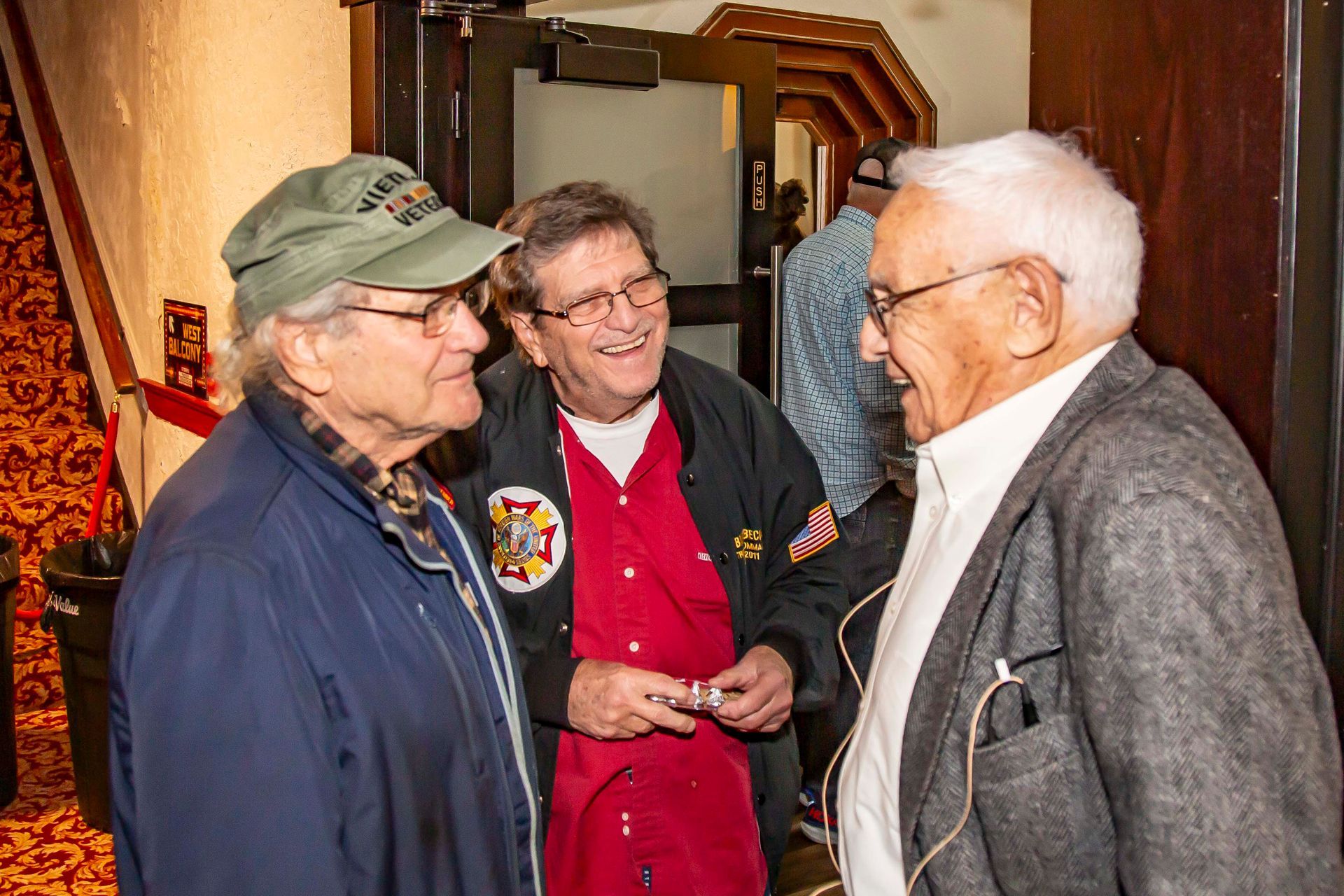 Three men conversing in a doorway; one wearing a green cap, another a red shirt, and the third a gray jacket.