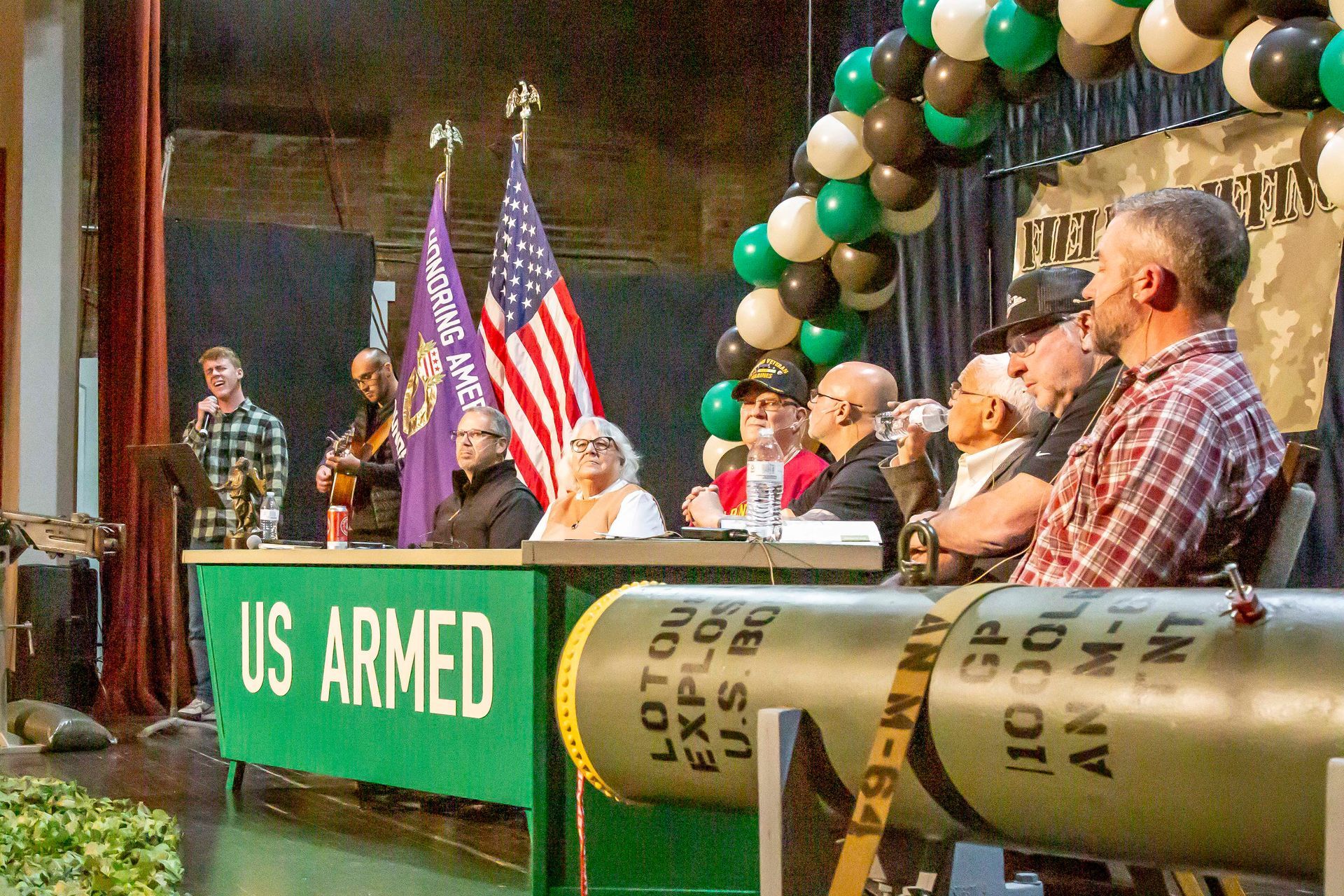Military appreciation event with speakers, US and purple flags, green table, and a large missile.