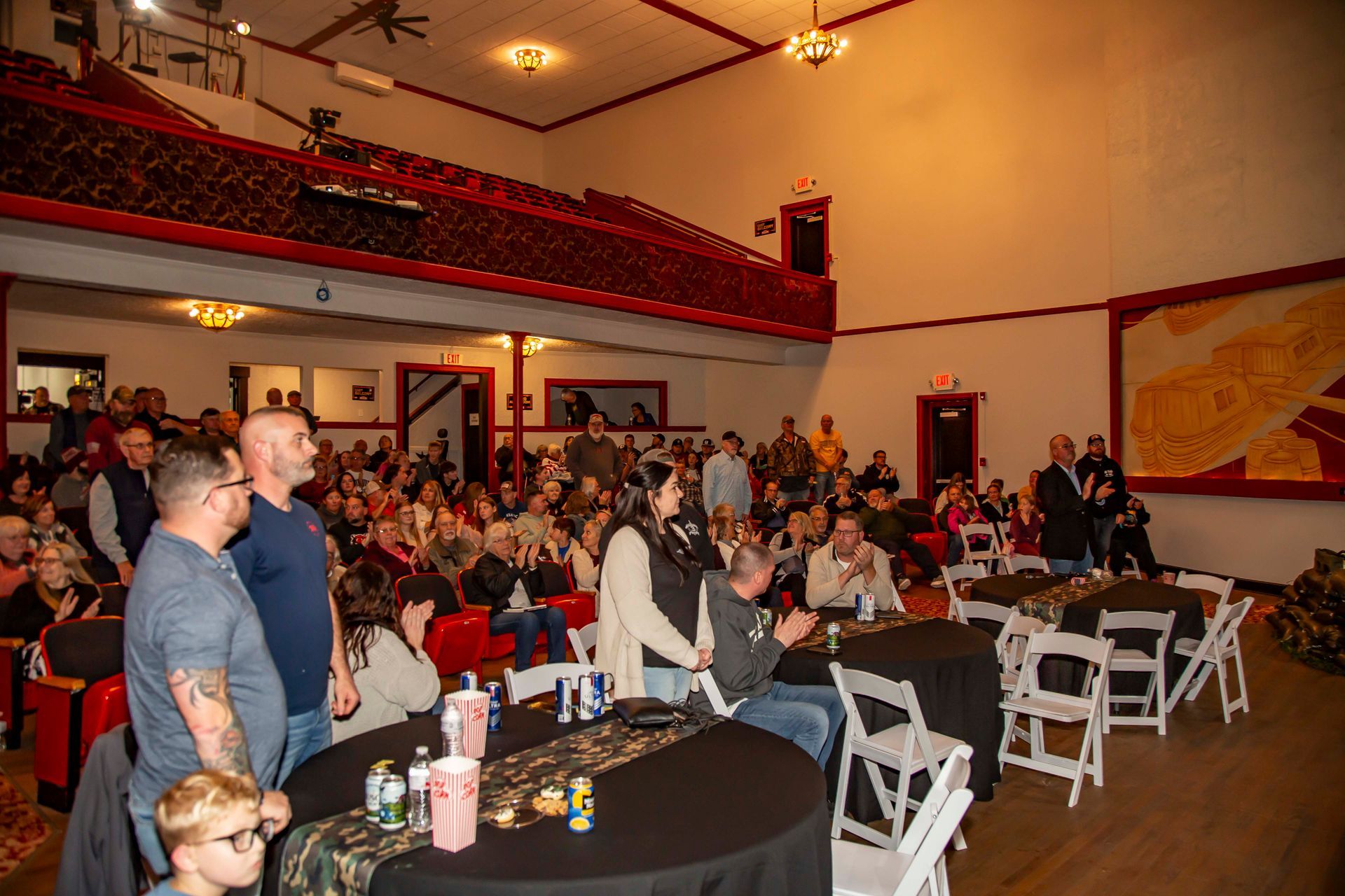 Audience in a theater with red accents, tables, and a balcony. People are watching or listening.