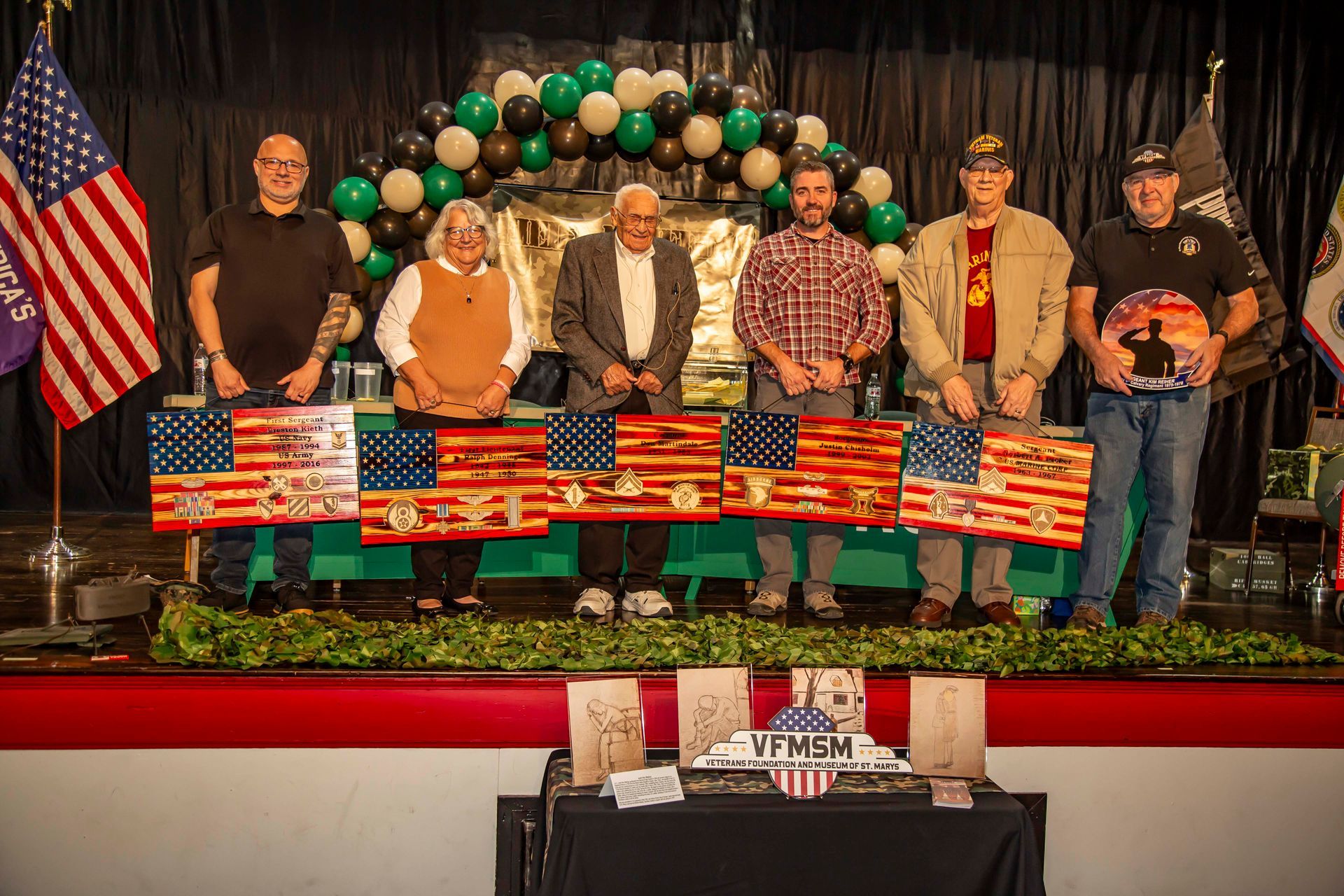 Six people stand on a stage holding American flag art, decorated with military themes. Green and black balloon arch in the background.