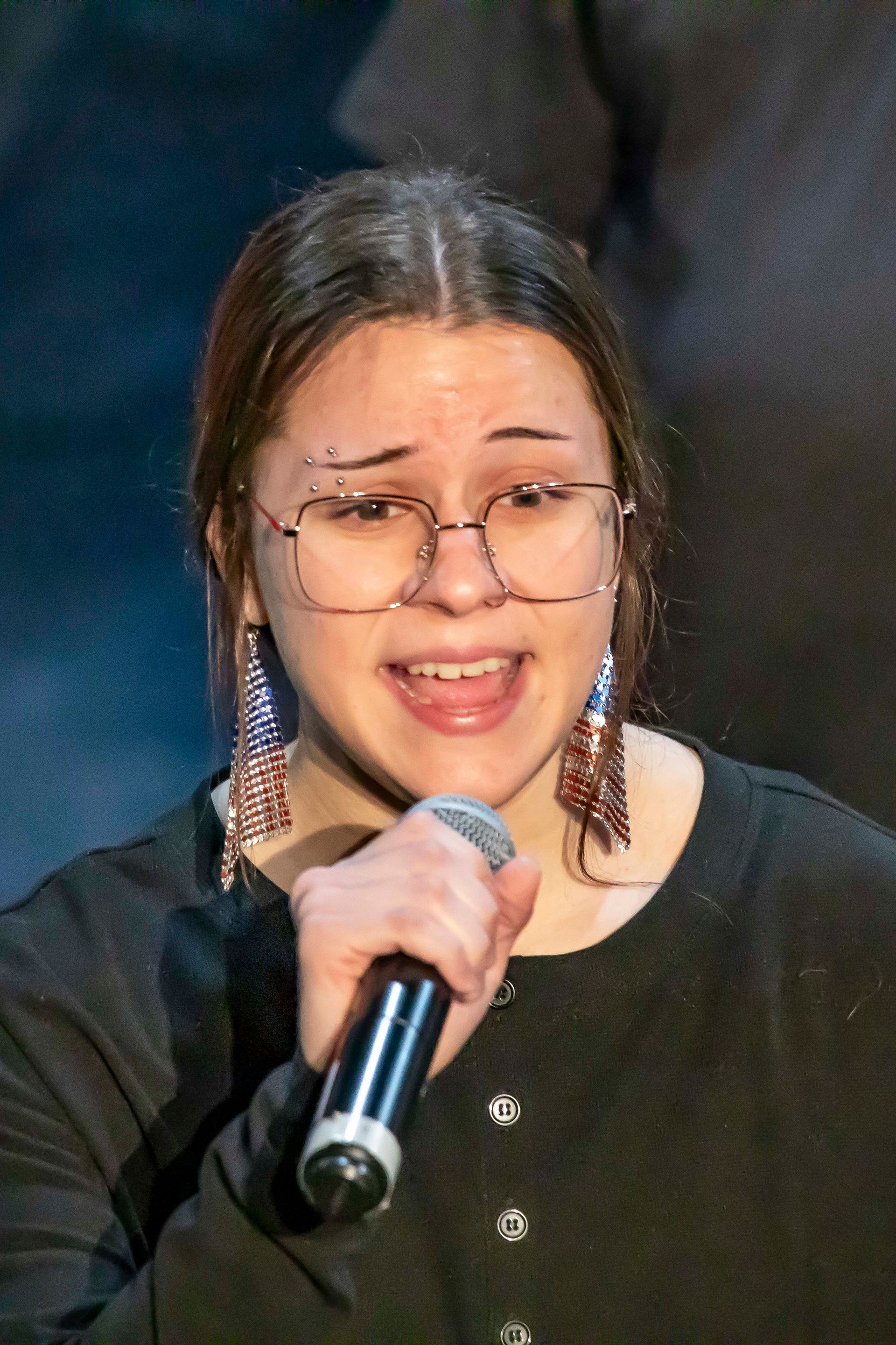 Person with glasses singing into a microphone, wearing long earrings, in front of a dark backdrop.