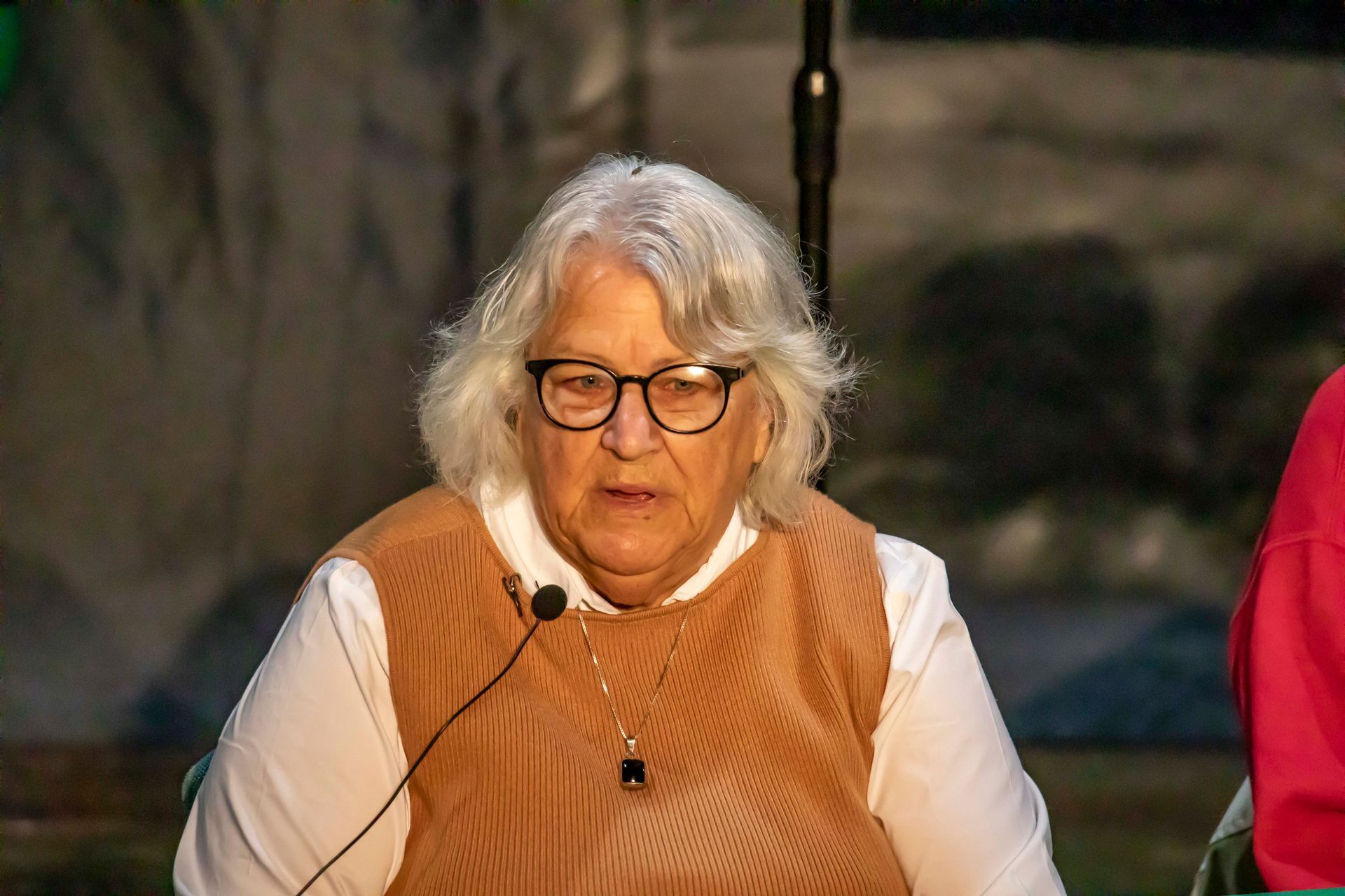 Woman wearing glasses and microphone, speaking at a public event.
