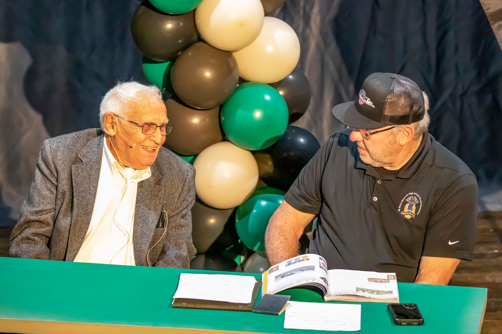 Two men sit at a table. One in a blazer, the other in a hat. Balloons in green, black, and white are in the background.
