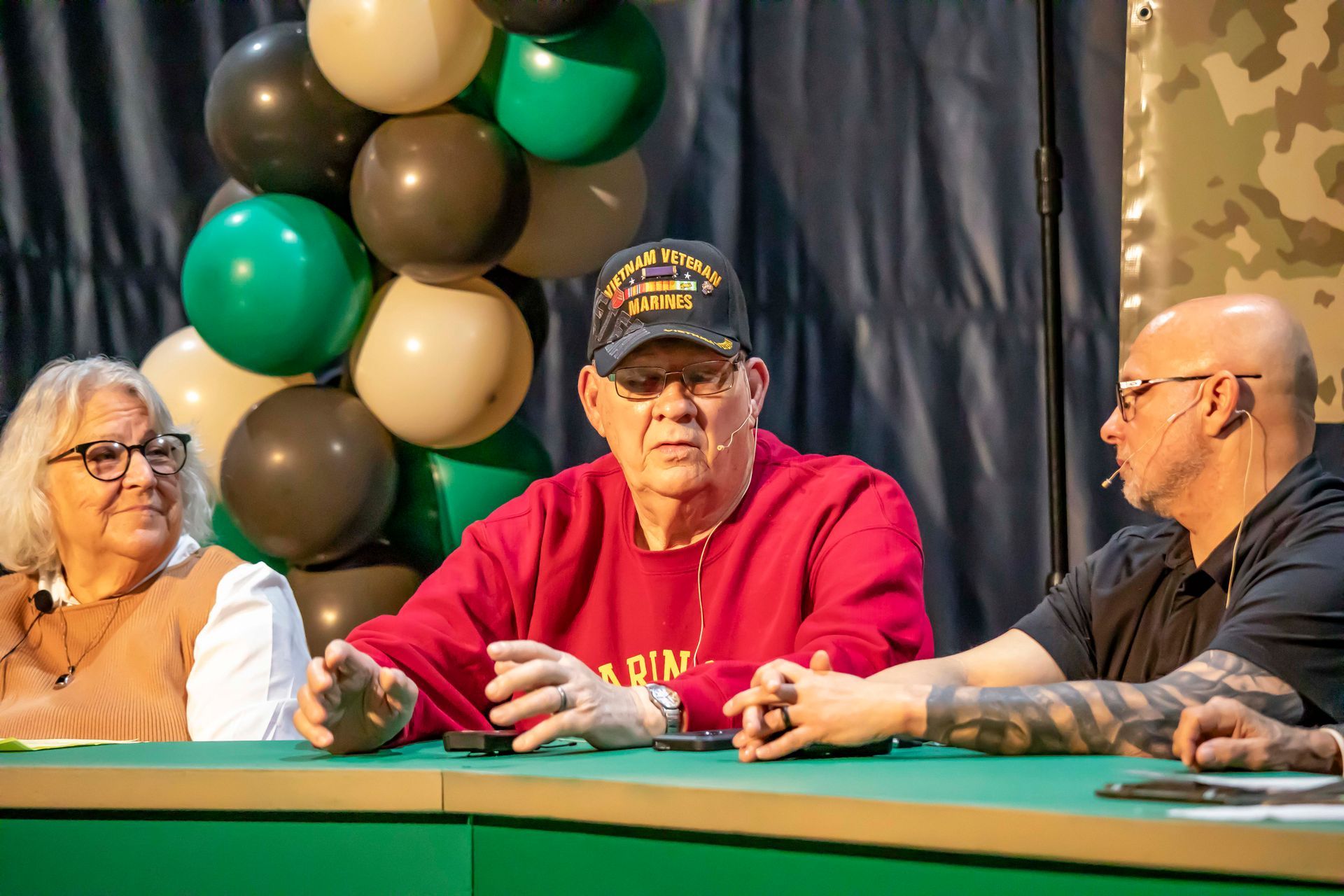 Three people seated at a table with a balloon arch in the background. Man in red sweater and hat speaking.