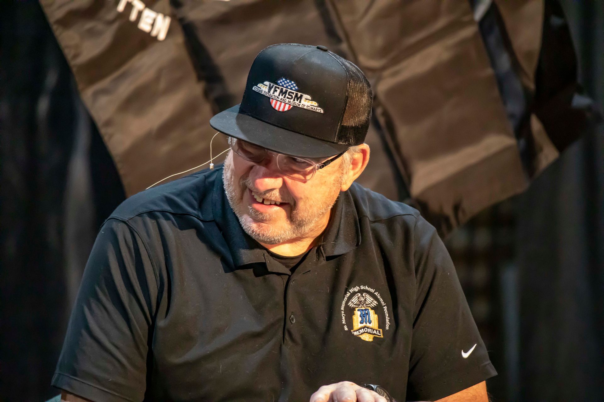Man wearing a hat and black polo shirt, sitting at a table and speaking.