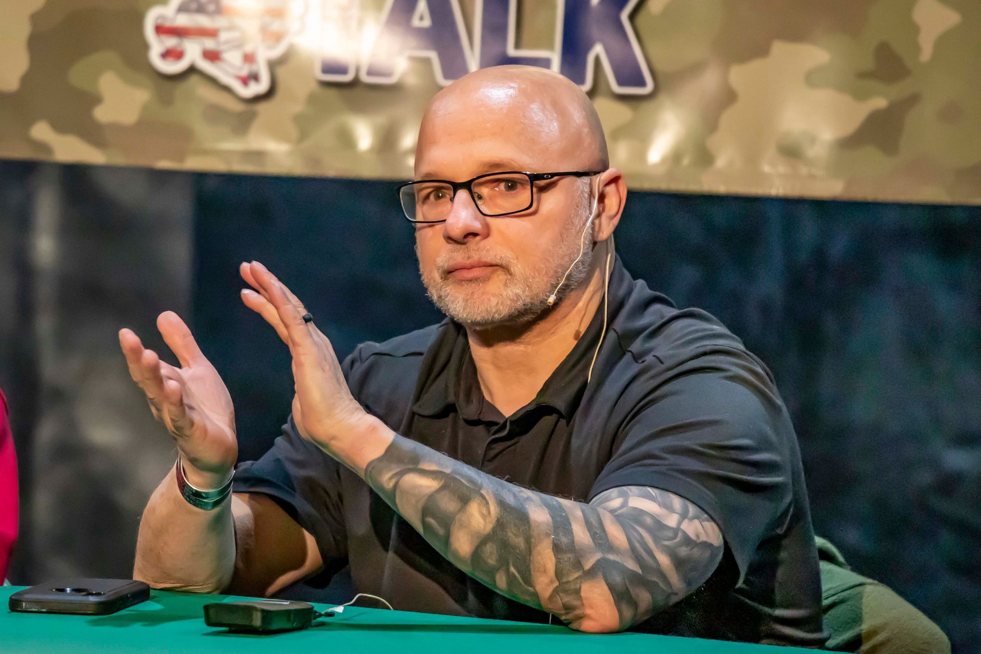 Man with glasses and arm tattoos gestures while speaking at a table with microphones.
