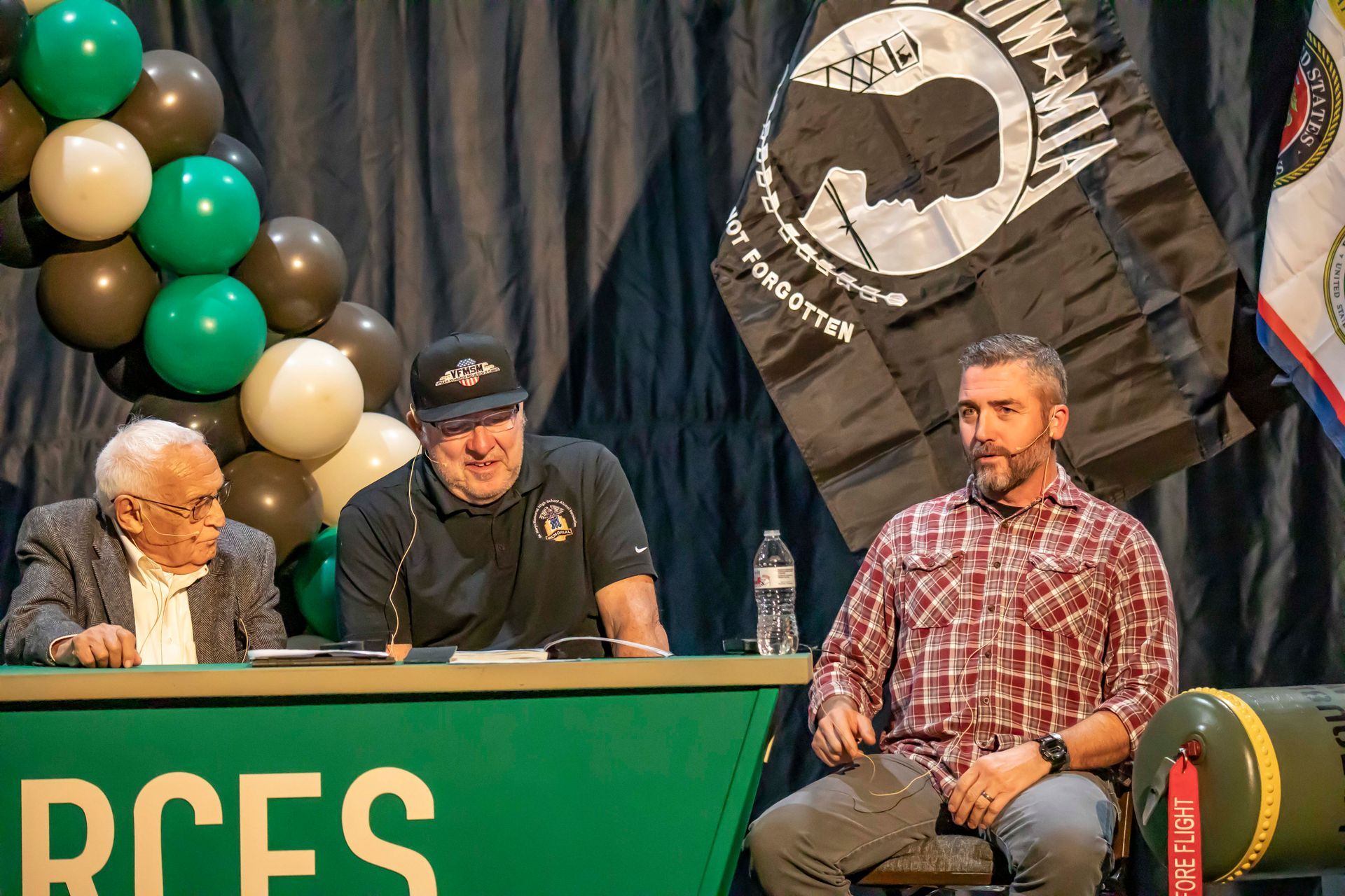 Three men at a table, one in plaid shirt speaking, others seated. POW-MIA flag in background, balloons.