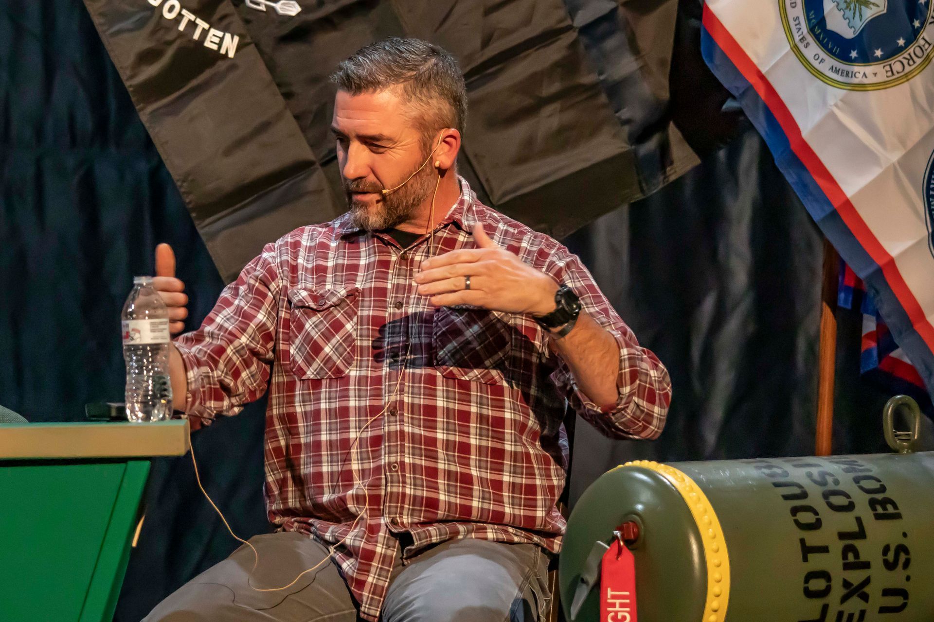 Man in plaid shirt speaking, gesturing. Sitting at a table, water bottle present. Military paraphernalia in background.