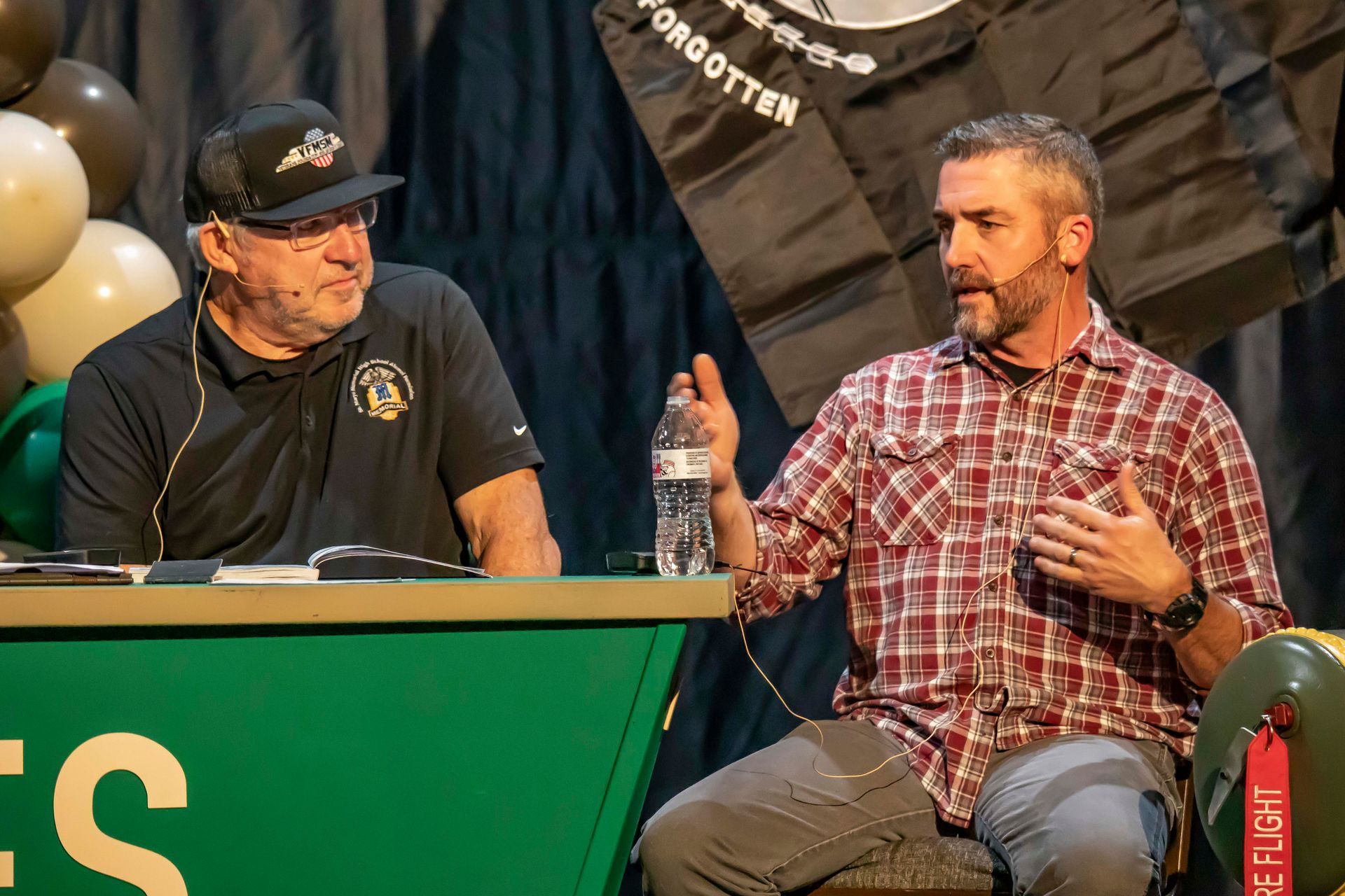 Two men seated at a table, one in plaid shirt speaking, the other listening. Stage backdrop with balloons and a sign.