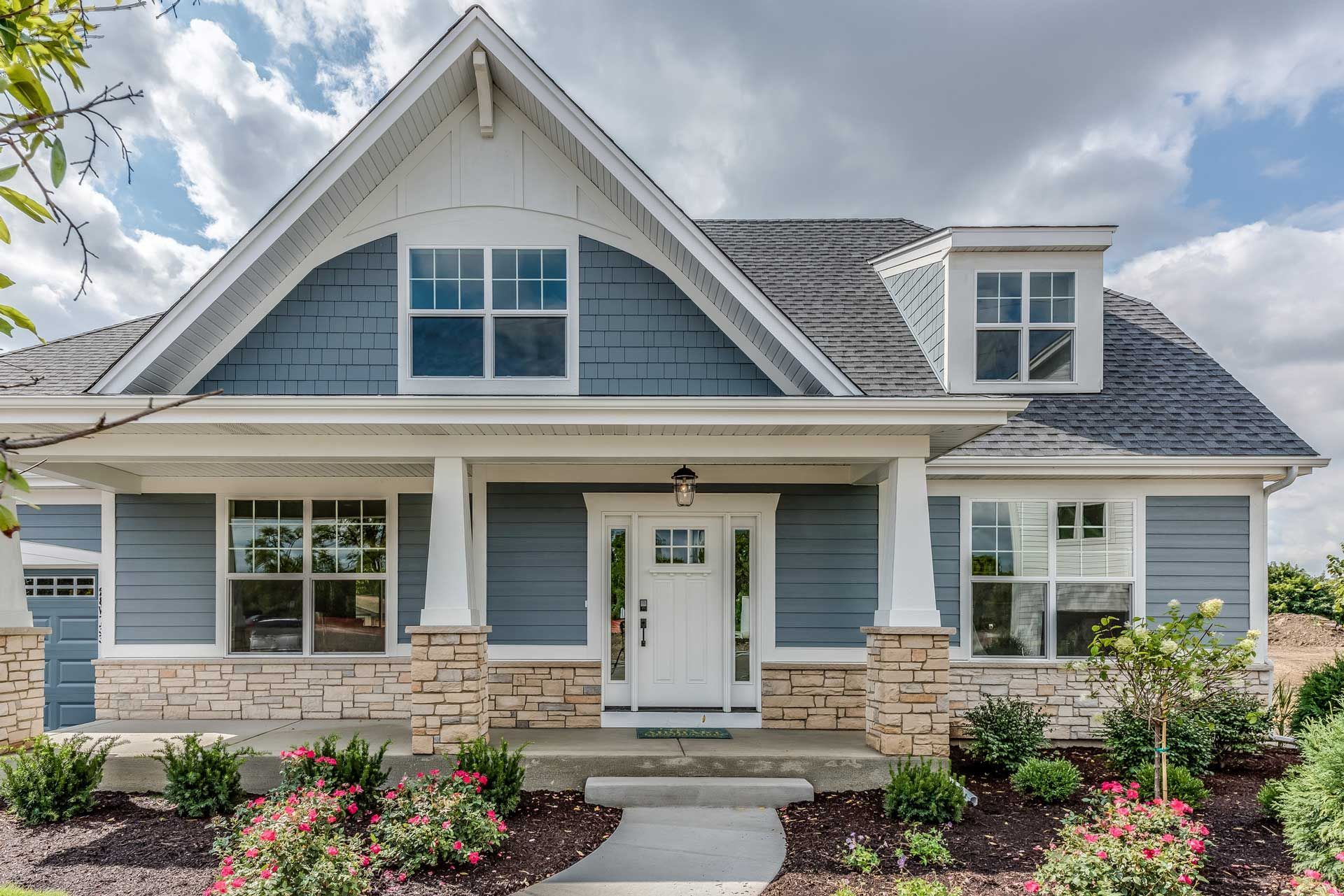 Blue and white Craftsman-style house with a stone base, front porch, and landscaping under a partly cloudy sky.