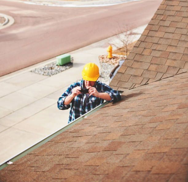 Construction worker on a brown shingled roof, wearing a yellow hard hat, near a sidewalk.