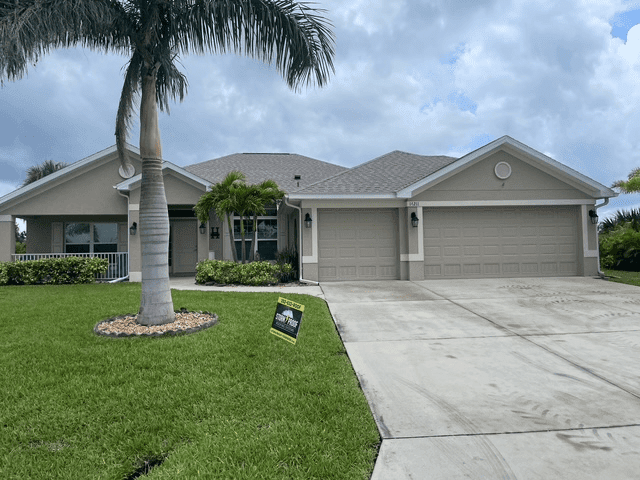 Tan house with gray roof, two-car garage, palm tree, and green lawn under a cloudy sky.