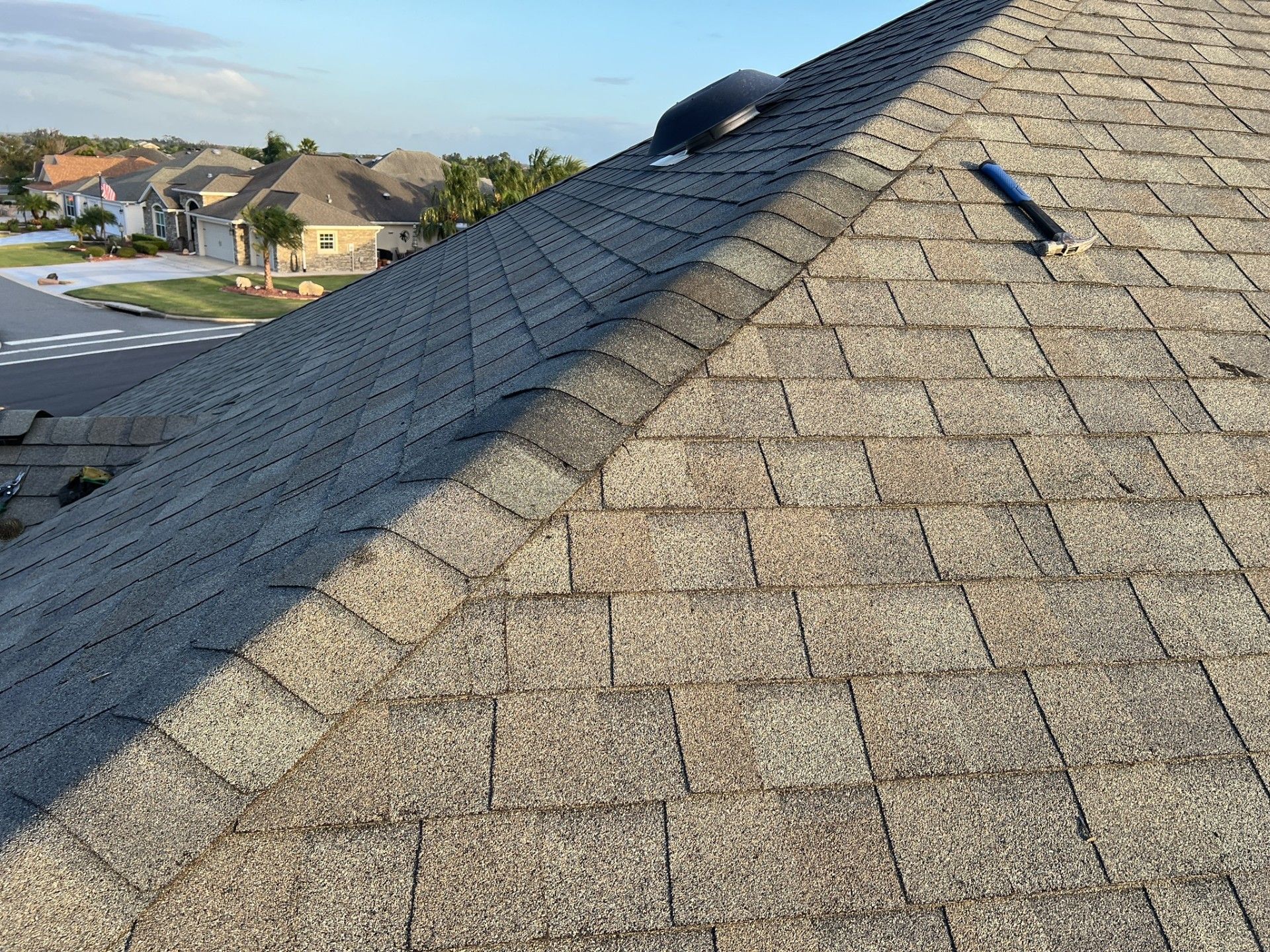 A shingled roof with a vent pipe and ridge cap, angled view over a suburban neighborhood.