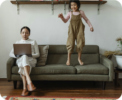 A woman is sitting on a couch with a laptop and a child is jumping on the couch.