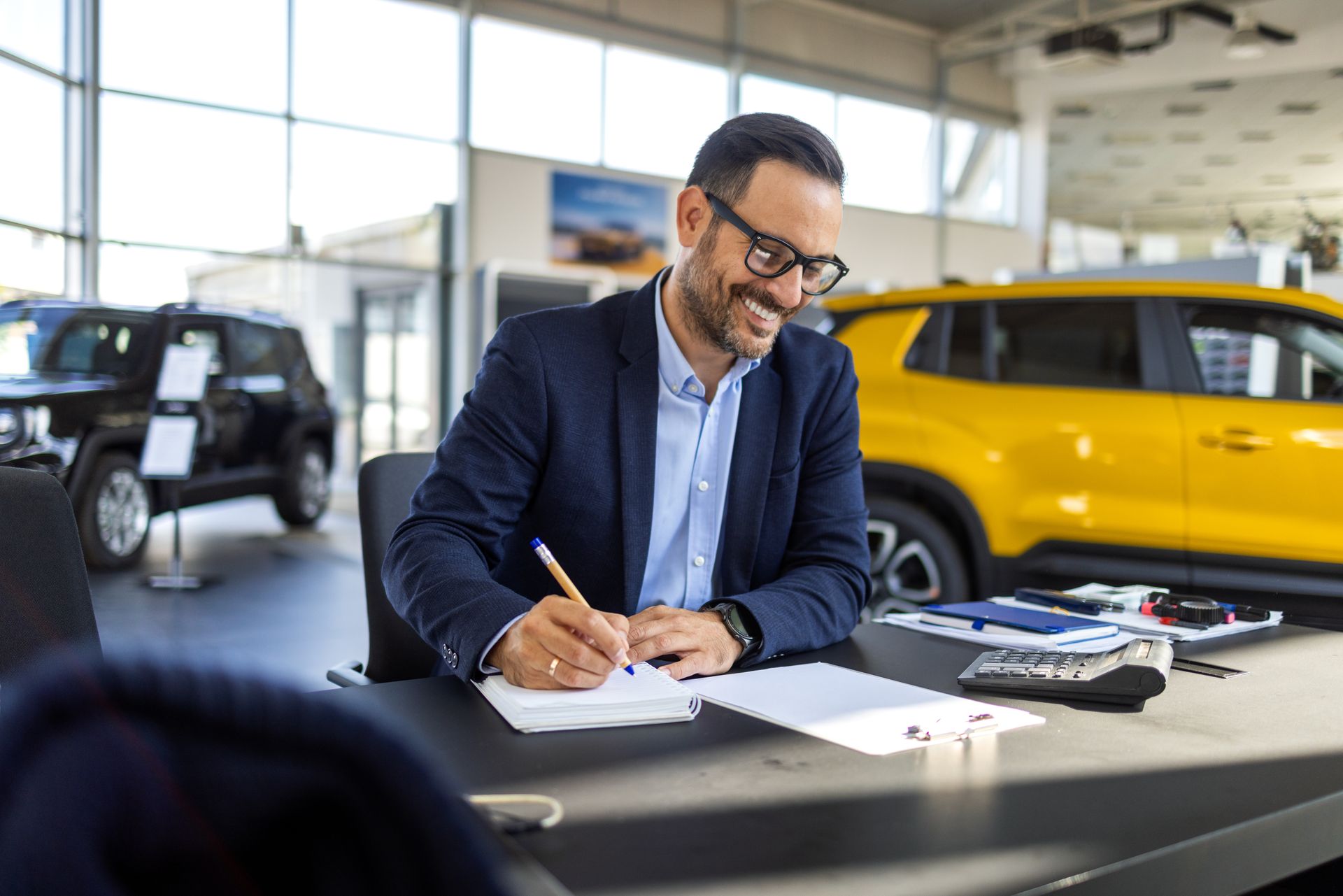 Man in glasses smiles, writing at a desk in a car dealership, a yellow car visible.