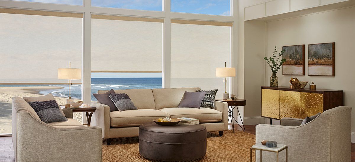 A living room with a couch , chairs , ottoman , and large windows overlooking the ocean.