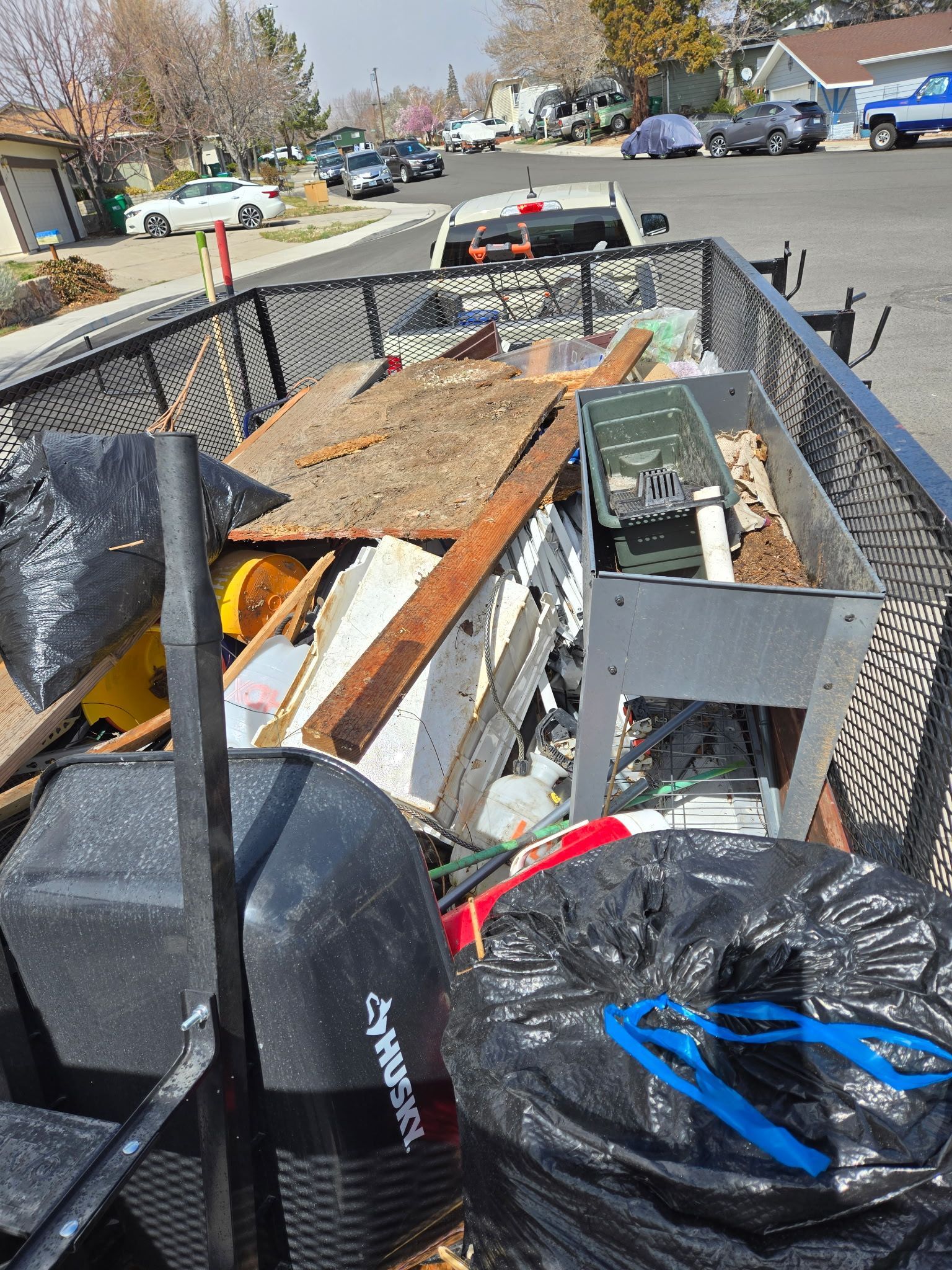 A trailer filled with trash and debris, including lumber and bags, parked on a residential street.