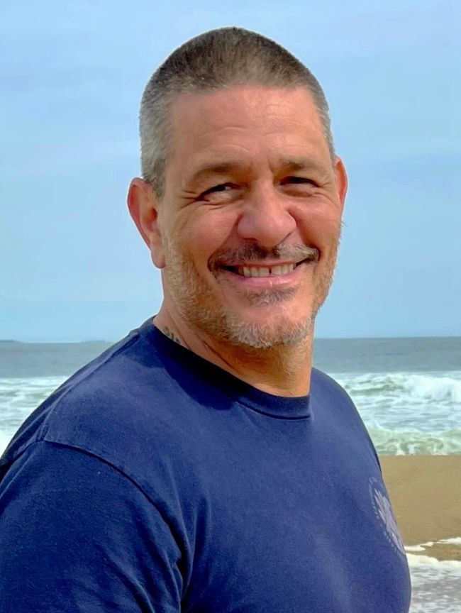 Man with short, graying hair smiles at camera on beach, wearing blue shirt. Ocean in background.