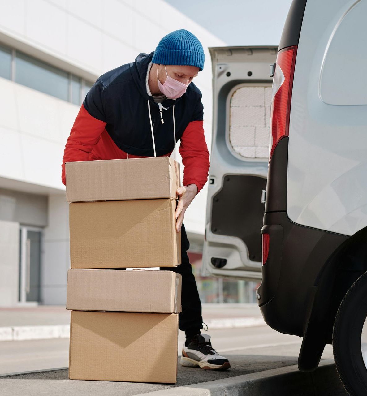 Delivery person in mask loading boxes from a van, wearing blue hat and red jacket, outdoors.