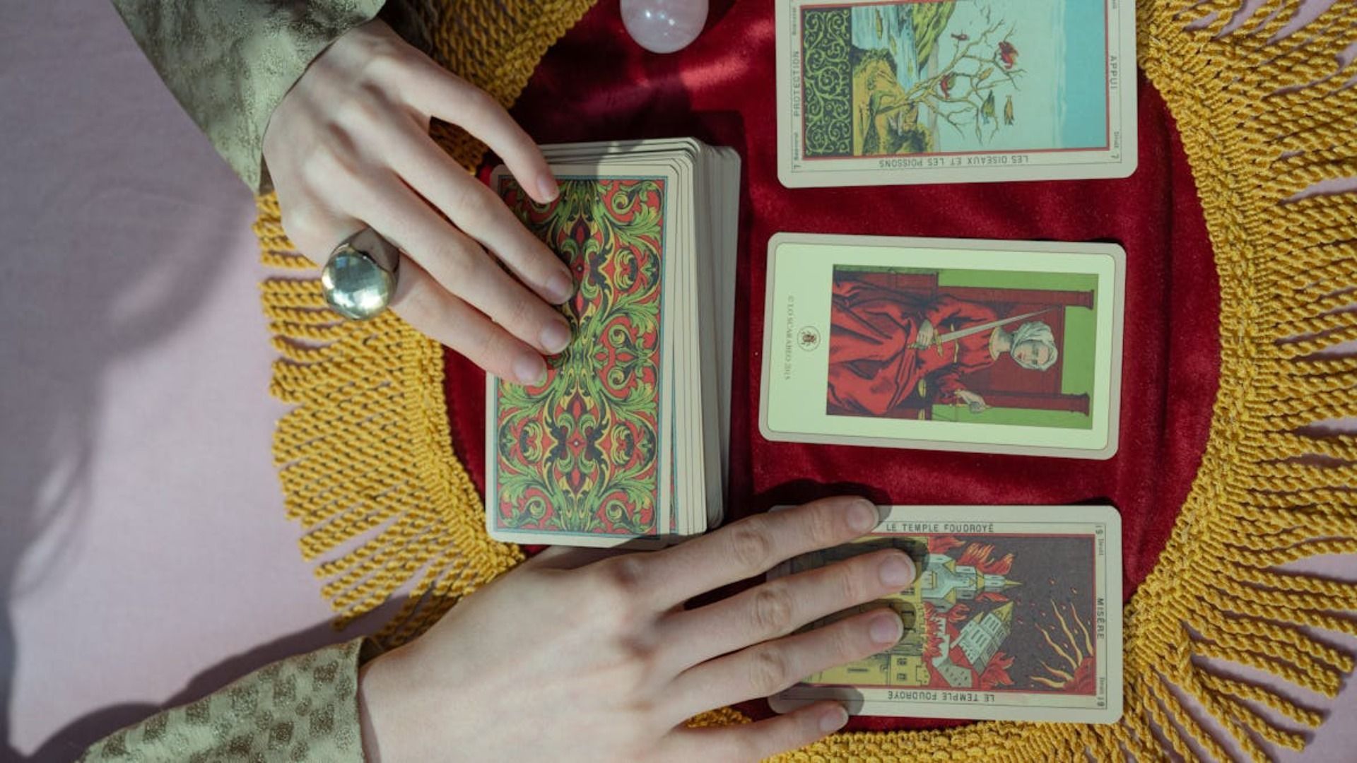 Manos barajando cartas del tarot sobre un paño de terciopelo con tres cartas boca arriba expuestas; se ve un cristal rosa.