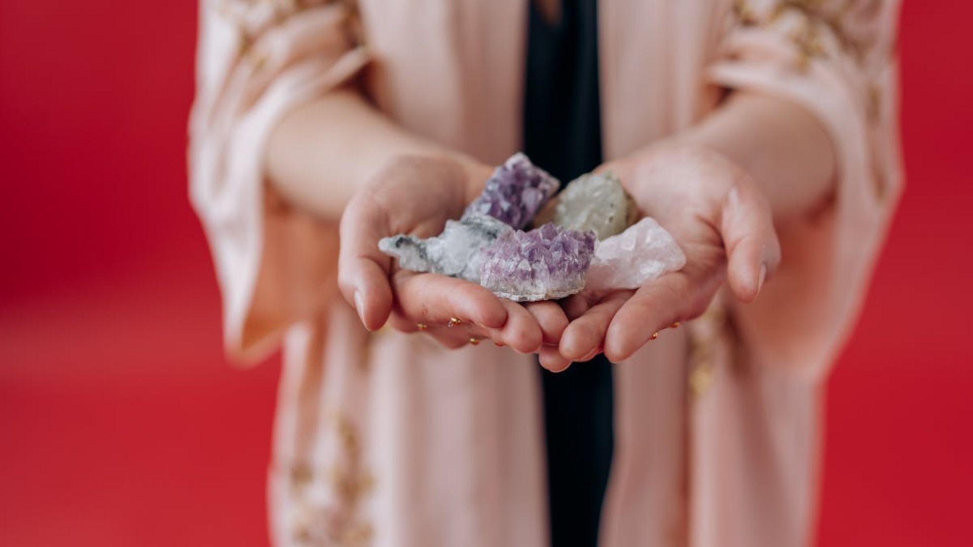 Manos sosteniendo un grupo de cristales de colores sobre un fondo rojo, vistiendo una túnica rosa.