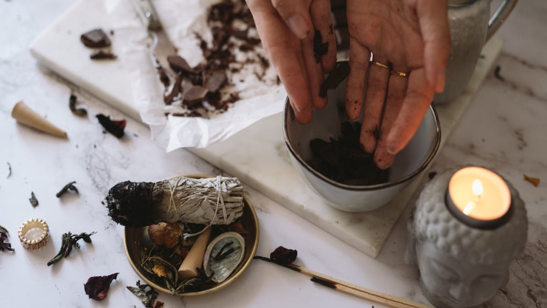 Manos preparando ingredientes en un tazón. Un manojo de salvia, una vela y otros objetos sobre una superficie de mármol.