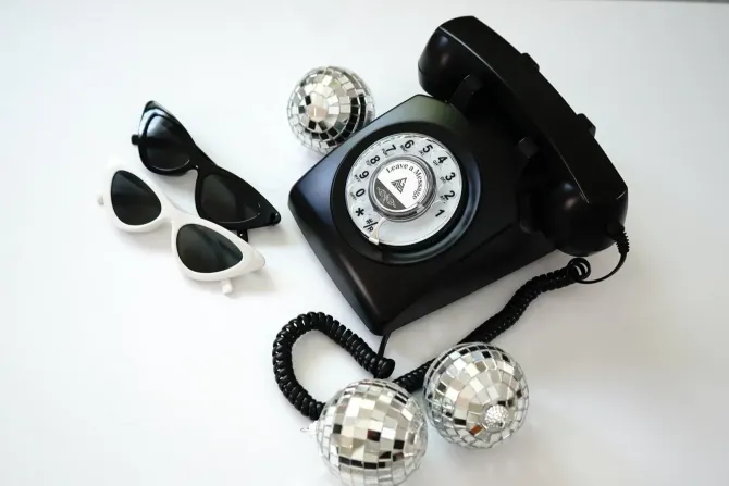 Black rotary phone, sunglasses, and disco balls on a white surface.