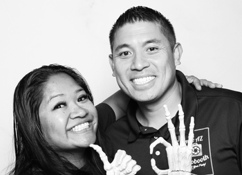 Two people smiling, posing in a photo booth. The person on the right has an arm around the other, holding a skeletal hand.