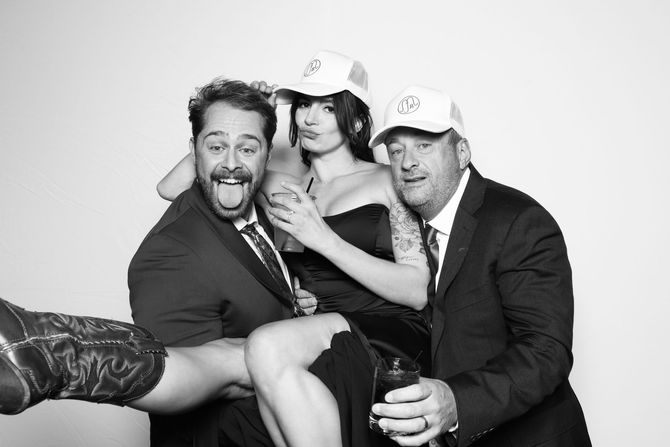 Three people pose in a photo booth: a woman held up by two men, all wearing branded trucker hats, with silly expressions.