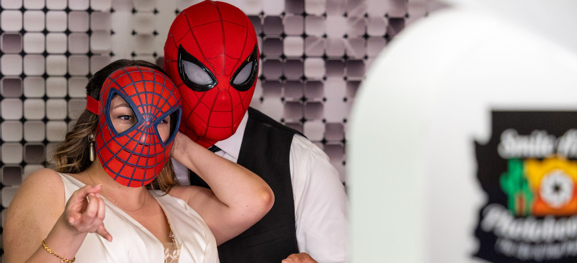Two people wearing Spider-Man masks posing for a photo. They are in front of a patterned backdrop.
