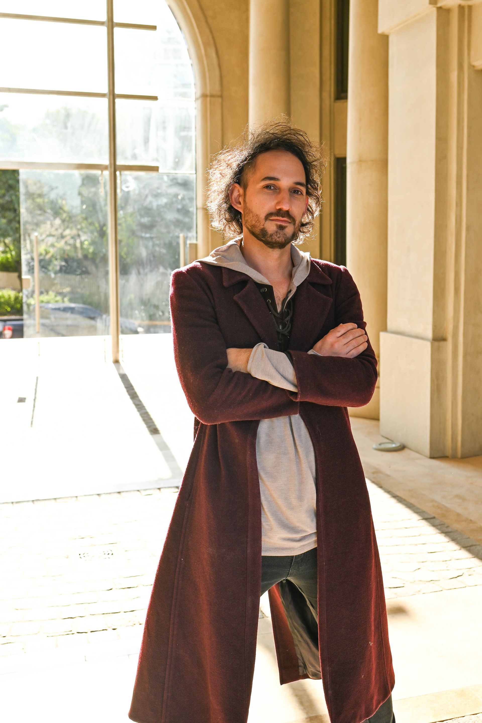 Hombre de pelo rizado con un abrigo granate, brazos cruzados, de pie en un pasillo soleado con columnas y grandes ventanas.